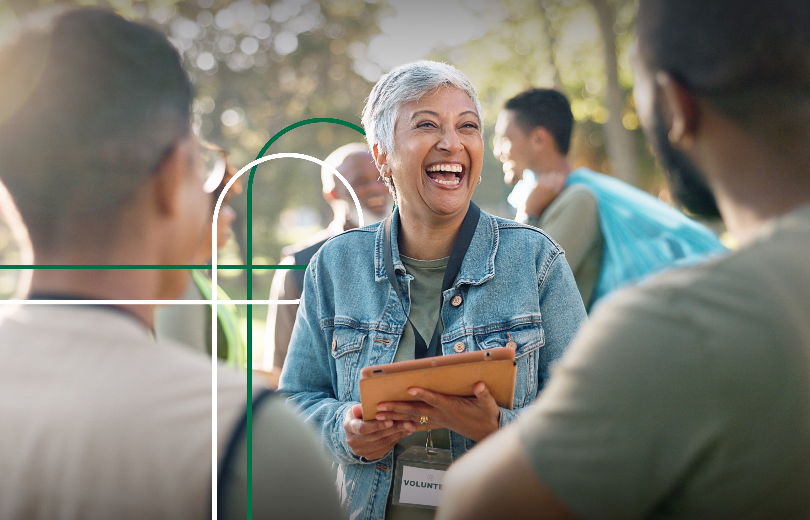 Alt text: A joyful older woman with short grey hair is laughing while holding a tablet, wearing a denim jacket and a "VOLUNTEER" badge. She is surrounded by other people in an outdoor setting, likely during a volunteer or community event. Sunlight filters through trees in the background.