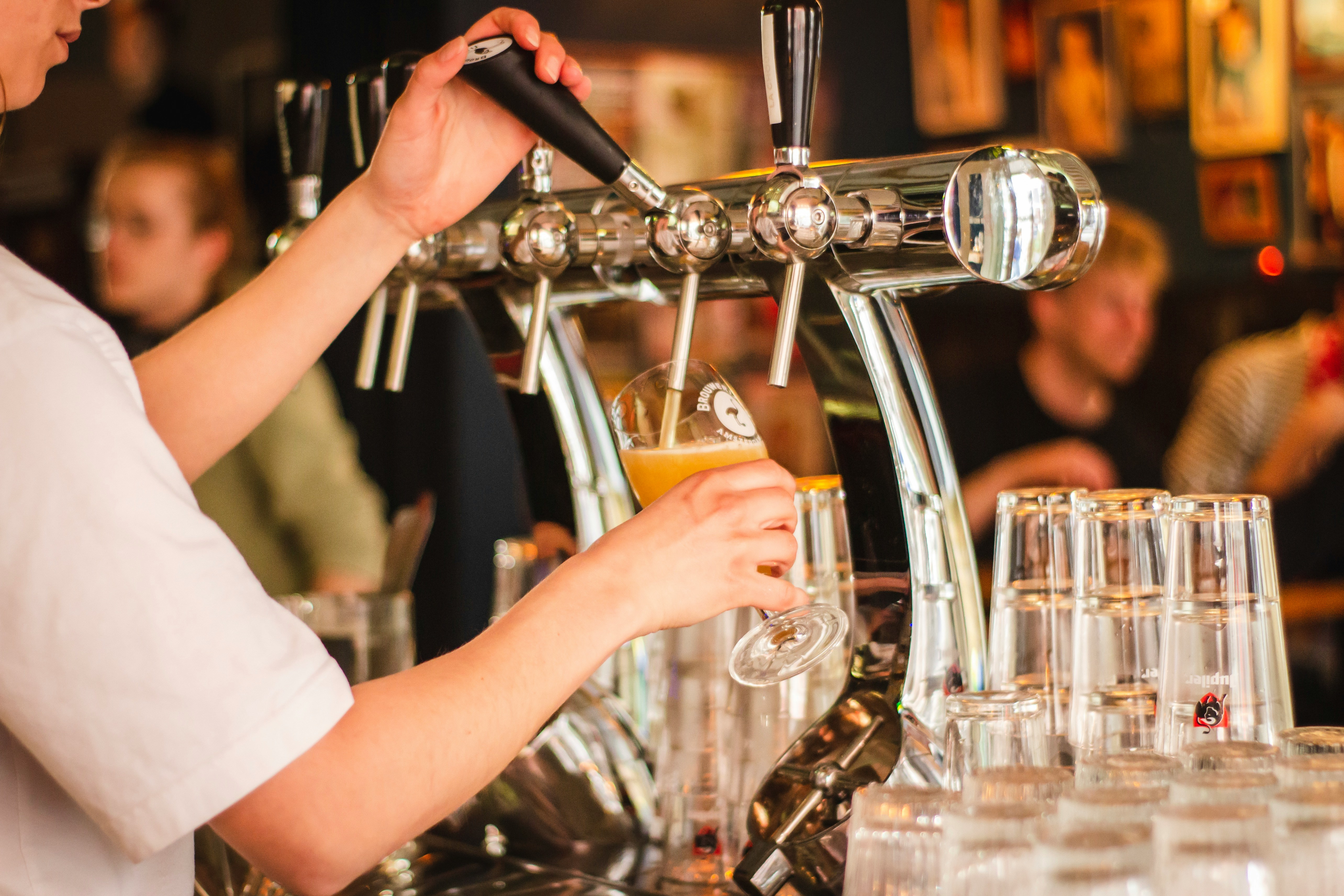 A bartender pours a pint of hazy craft beer from a chrome beer tap in a busy bar, with rows of clean glasses lined up and patrons blurred in the background.