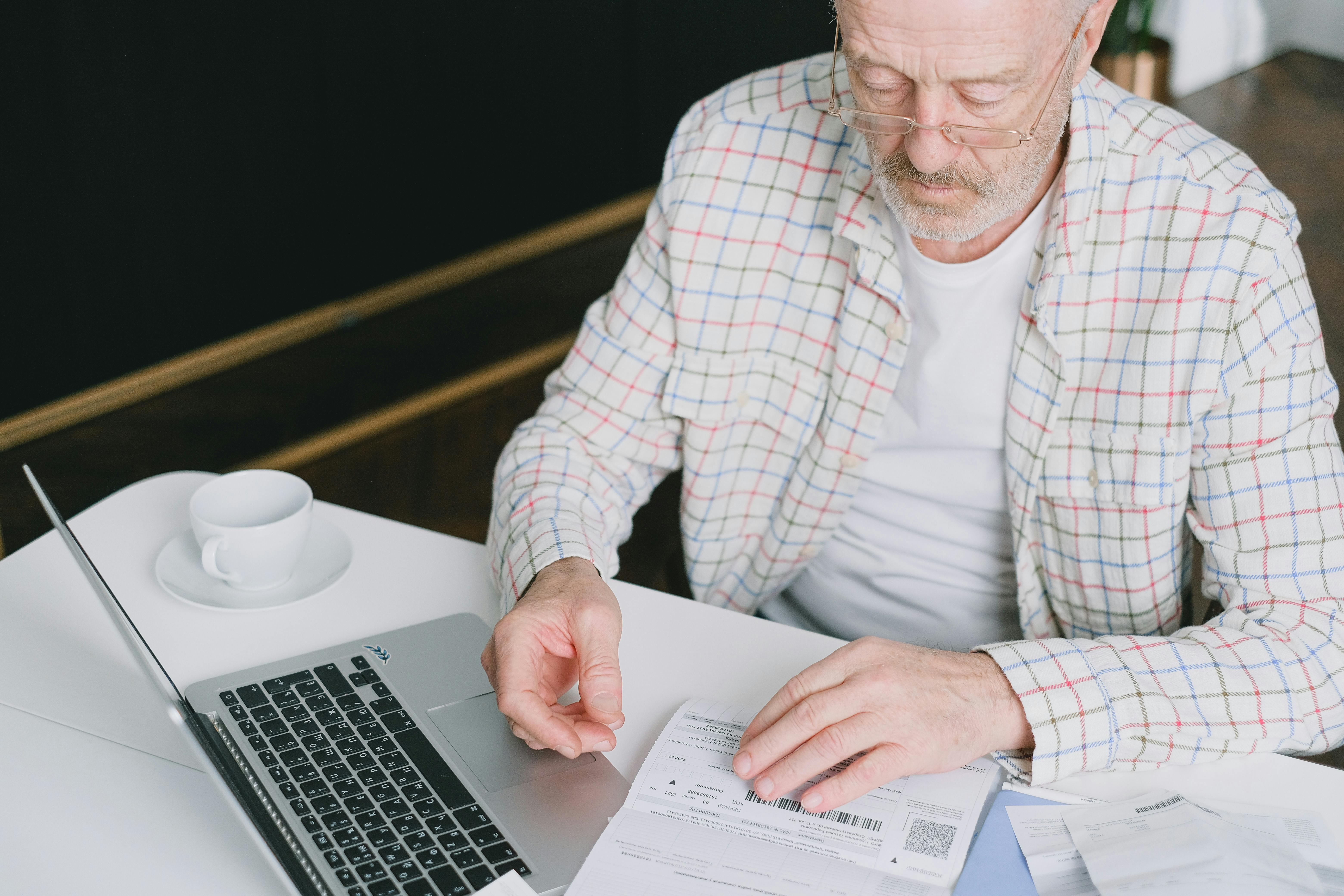 Older man wearing glasses and a plaid shirt sitting at a white table, reviewing documents next to an open laptop. A cup and saucer are placed nearby.
