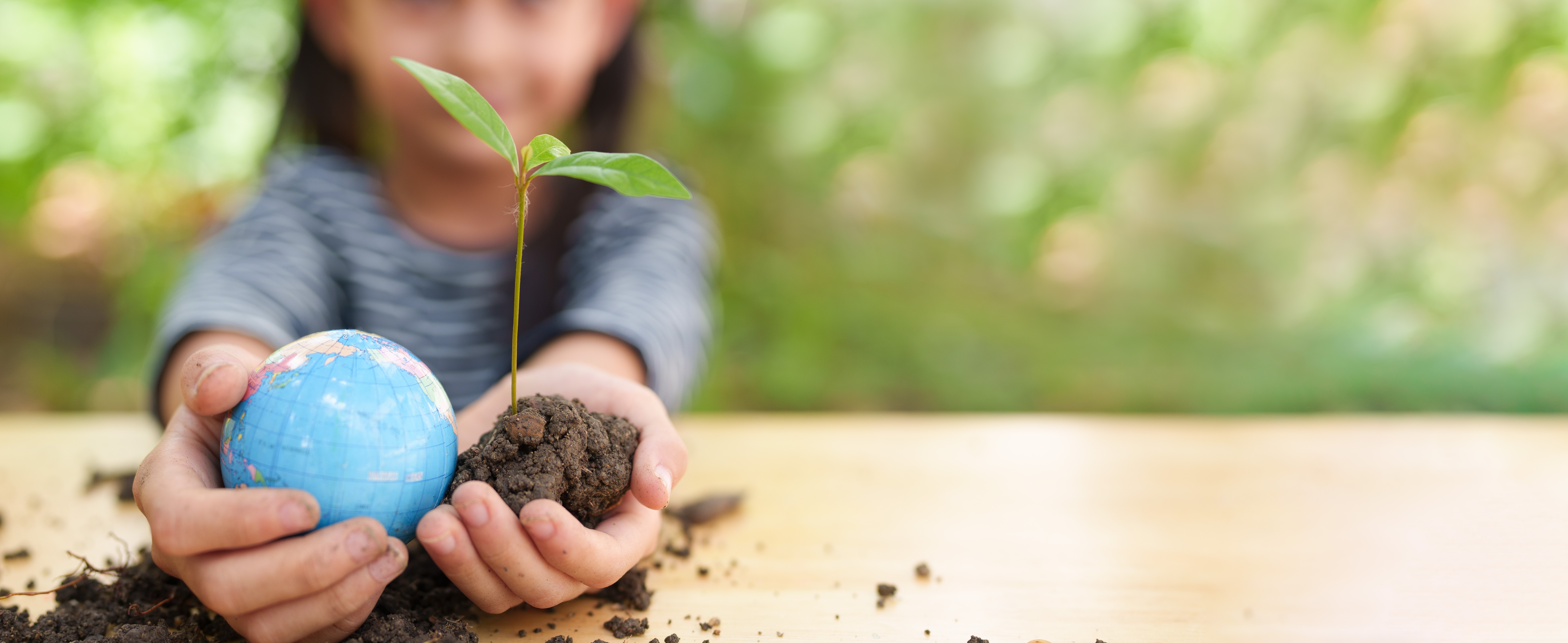 A child holds a small globe in one hand and a young green plant with soil in the other, symbolizing environmental protection and sustainability. The background is blurred greenery, creating a natural and hopeful atmosphere.