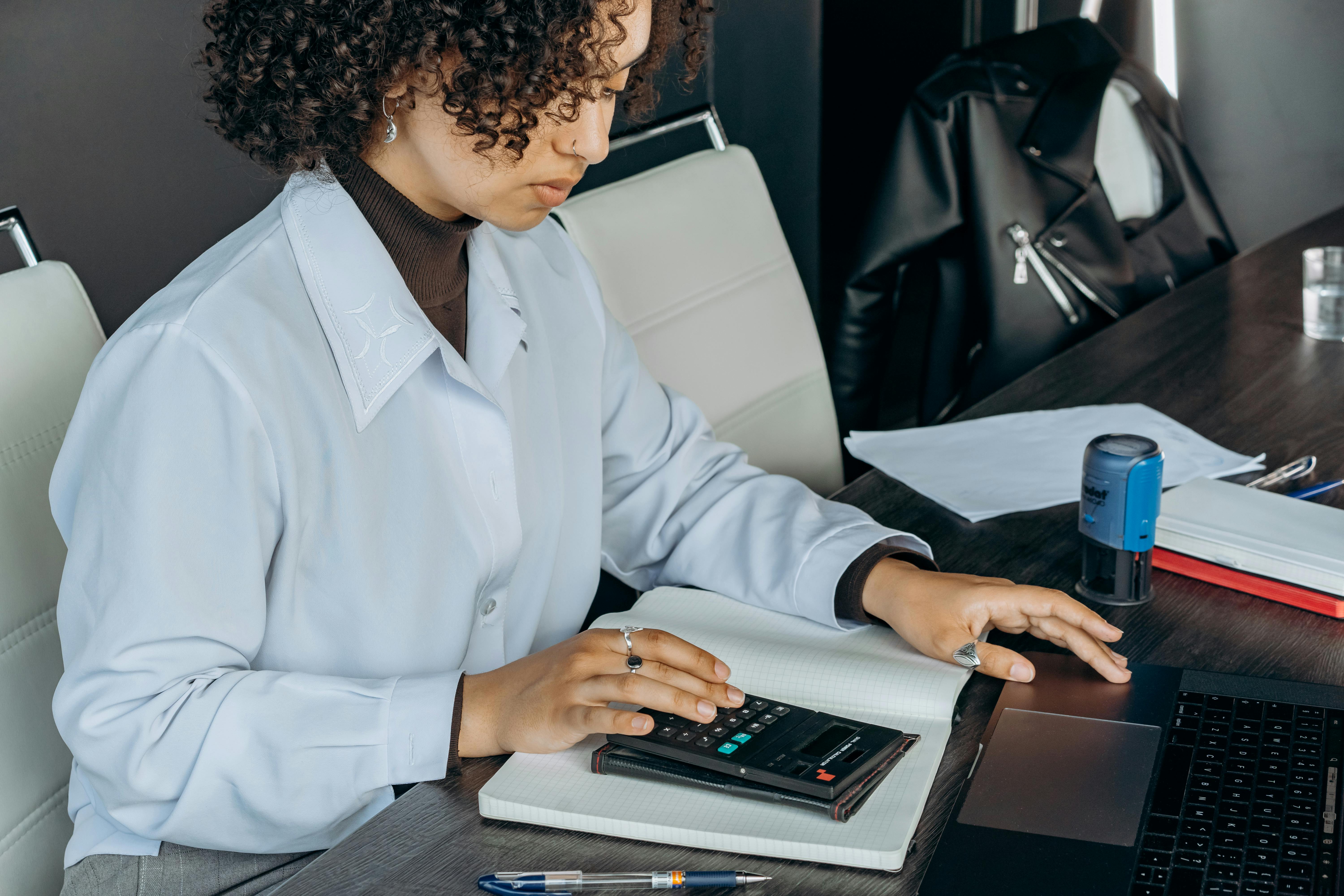 A woman with curly hair wearing a white blouse and dark turtleneck sits at a desk using a calculator and laptop. She is focused on her work, with papers, a notebook, a pen, and a blue self-inking stamp on the desk beside her.