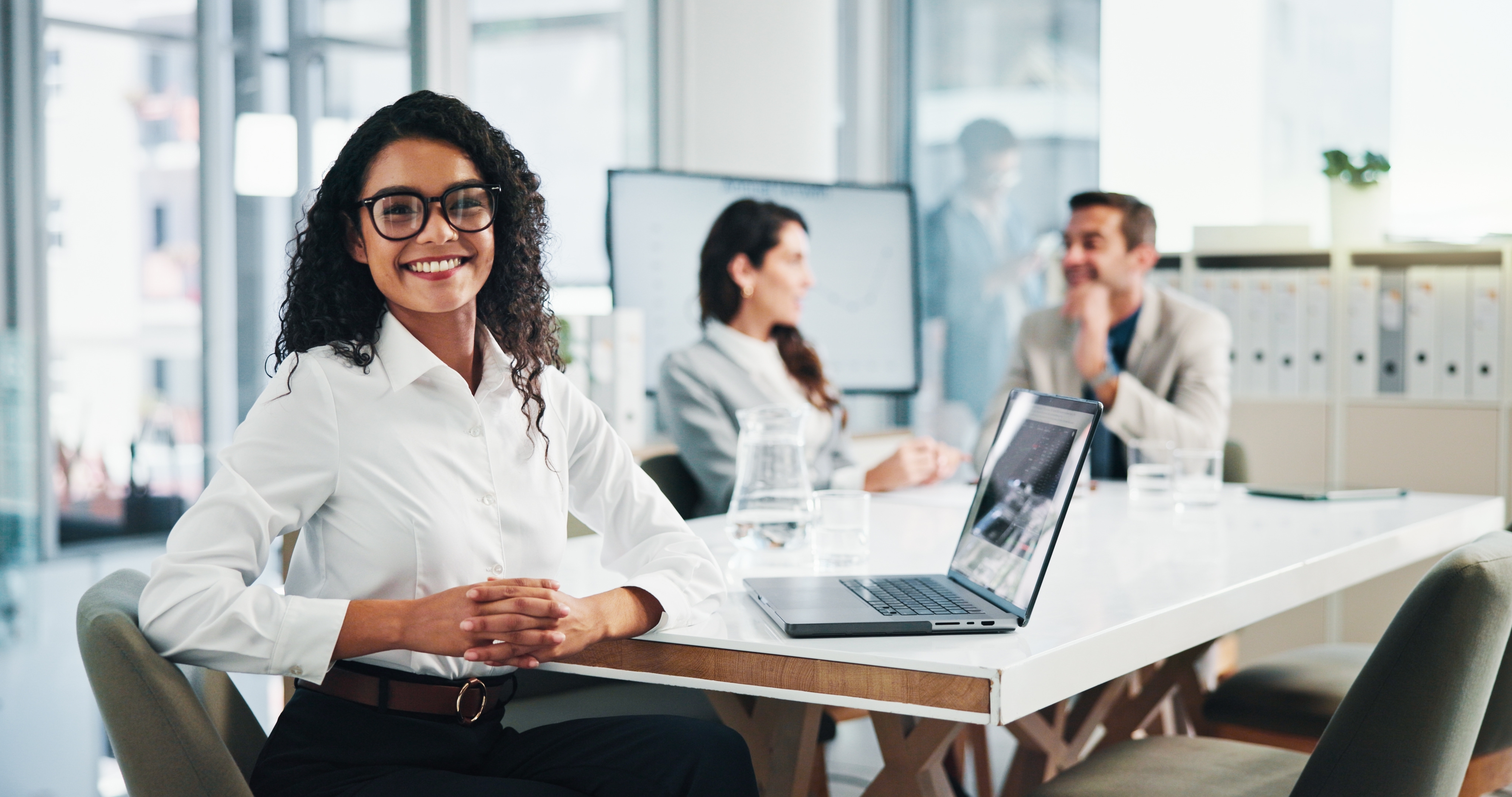 A woman sits at a desk with a laptop, smiling at the camera, while two colleagues have a discussion in the background in a modern office setting.