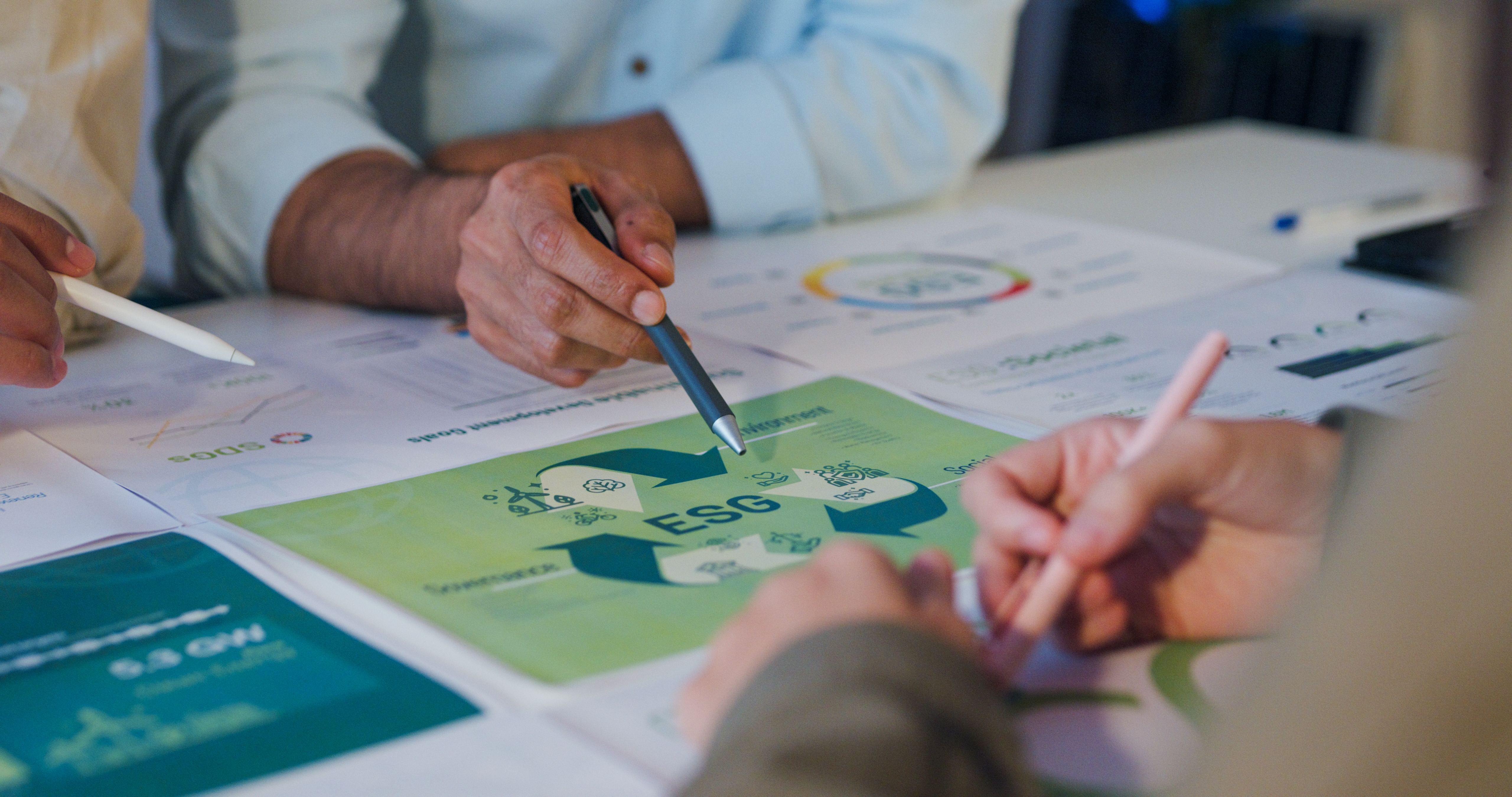 Close-up of people working on ESG (Environmental, Social, and Governance) reports, with hands holding pens and pointing at sustainability charts and documents on a desk.