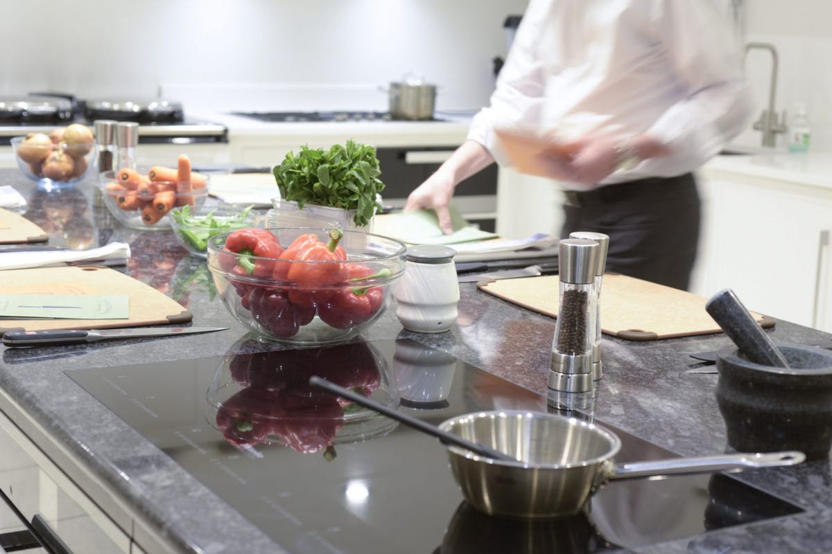 A modern kitchen workspace with fresh vegetables, herbs, and cooking utensils on a countertop, while a chef prepares food in the background.