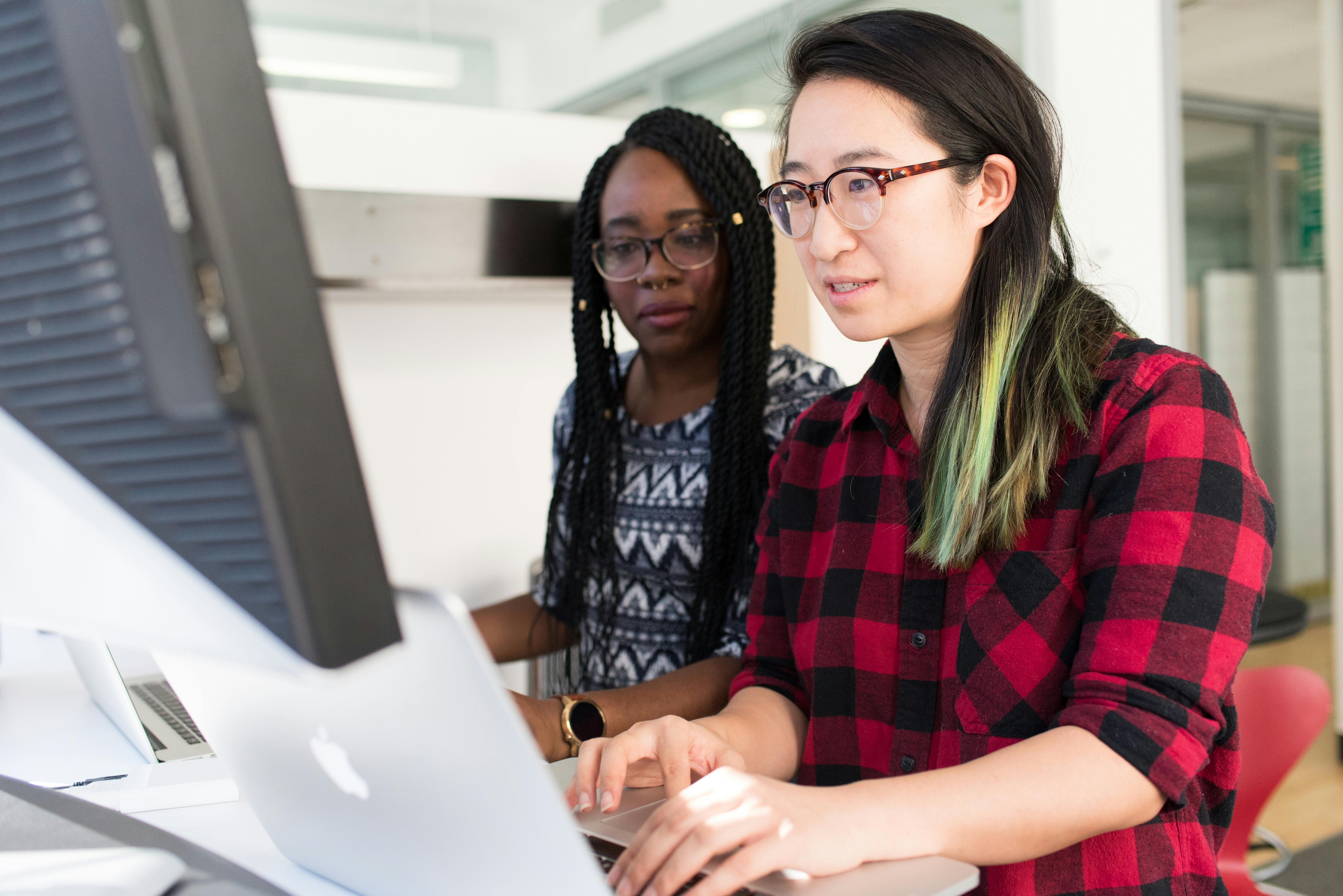 Two women working together at a desk, looking at a computer screen. One is typing on a laptop while the other observes, both focused on the task.