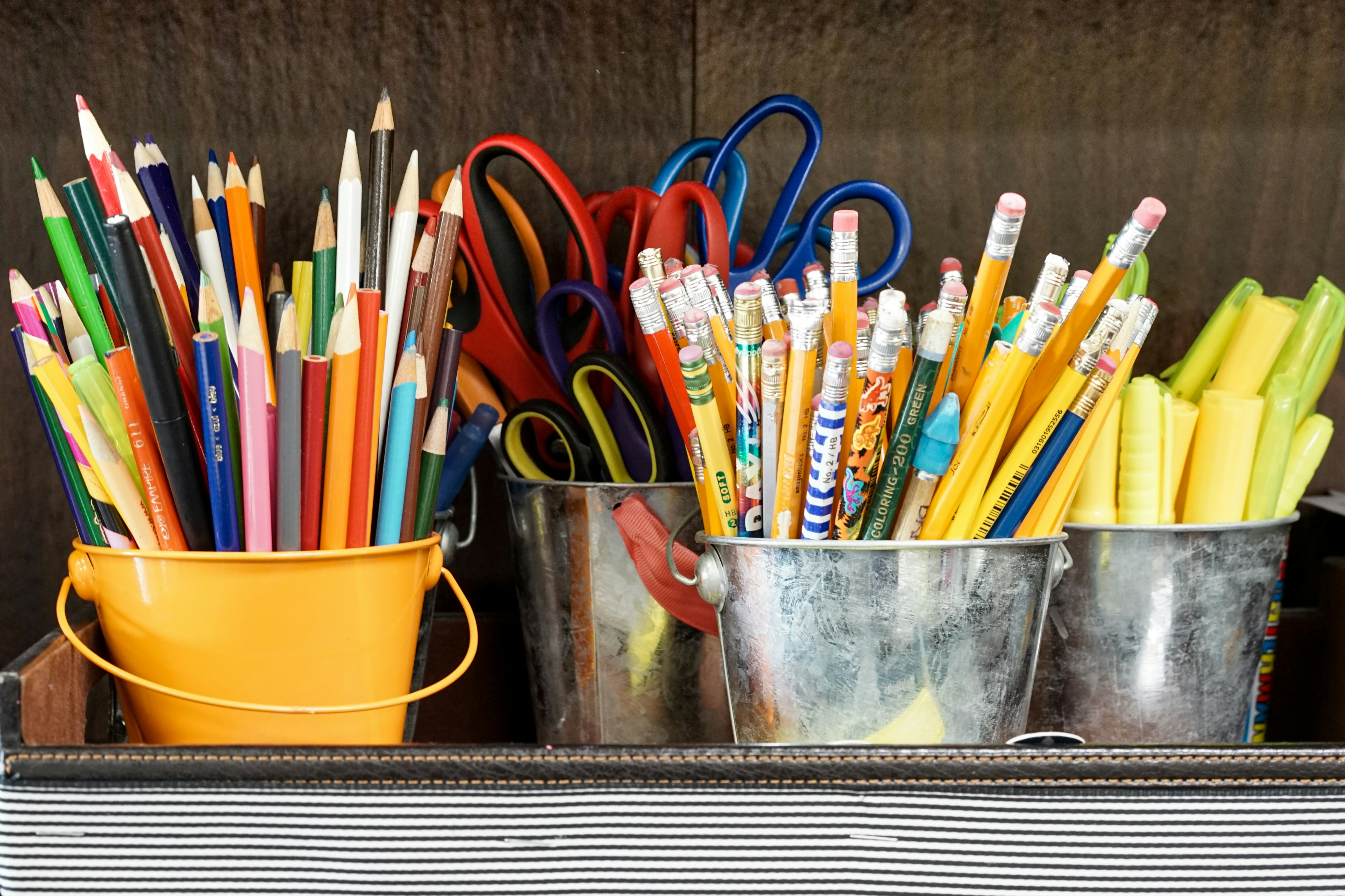 Assorted colorful pencils, scissors, and highlighters are organized in three metal buckets. The image conveys an atmosphere of creativity and order.