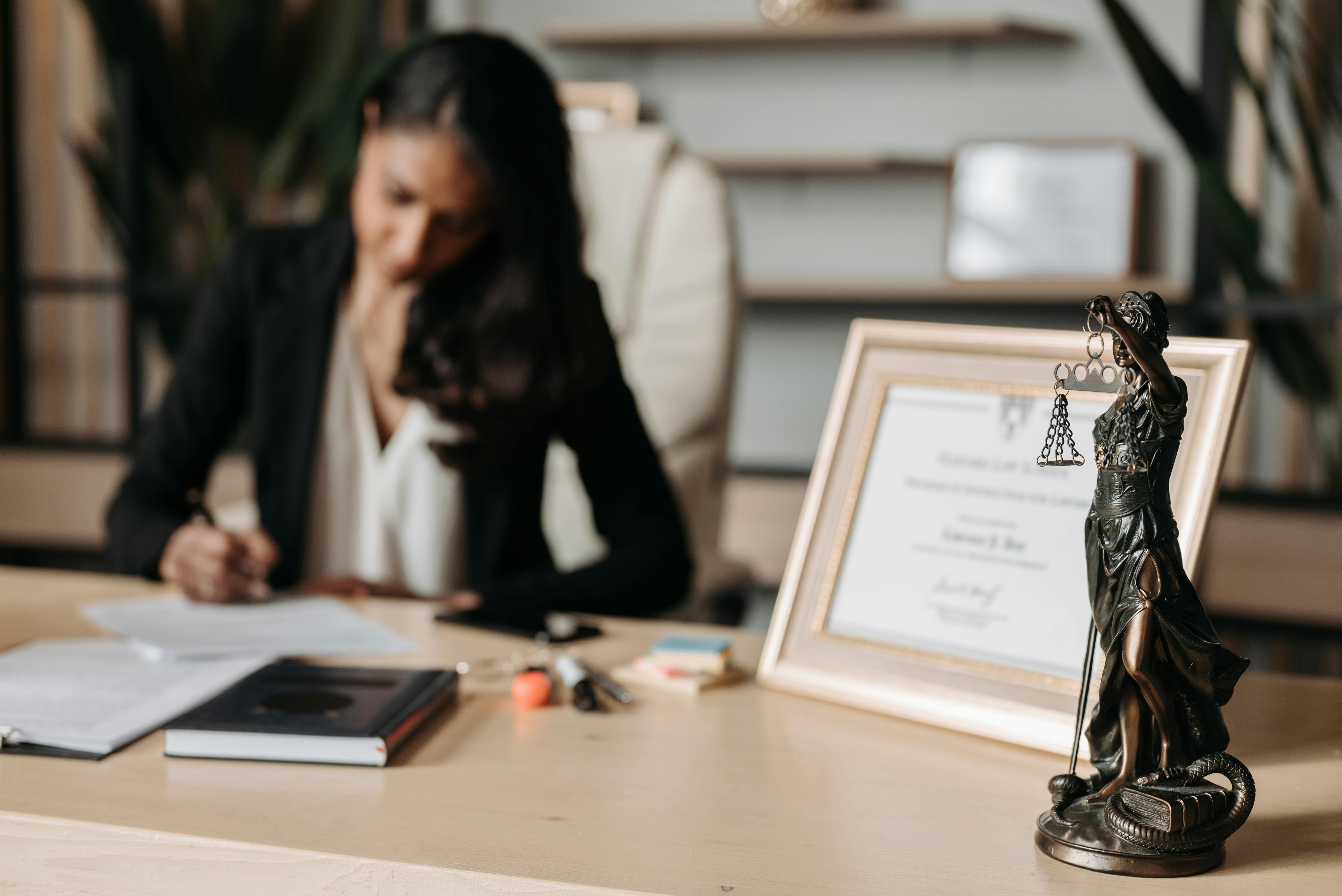 A woman in a black blazer writes at a desk with a "Lady Justice" statue and framed certificate in the foreground, conveying focus and professionalism.
