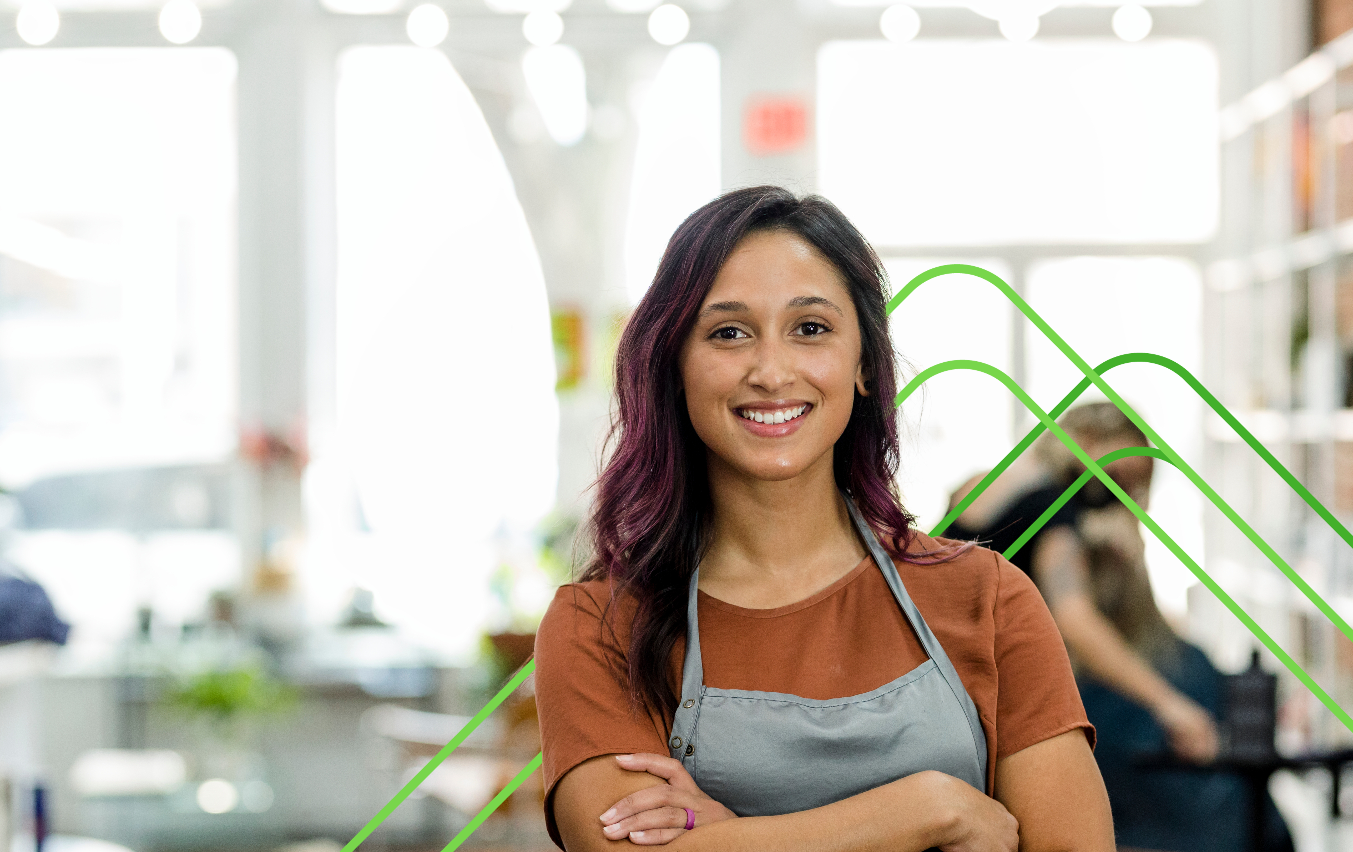 a smiling woman wearing a brown shirt and a grey apron, standing confidently with her arms crossed in a bright, modern workspace.