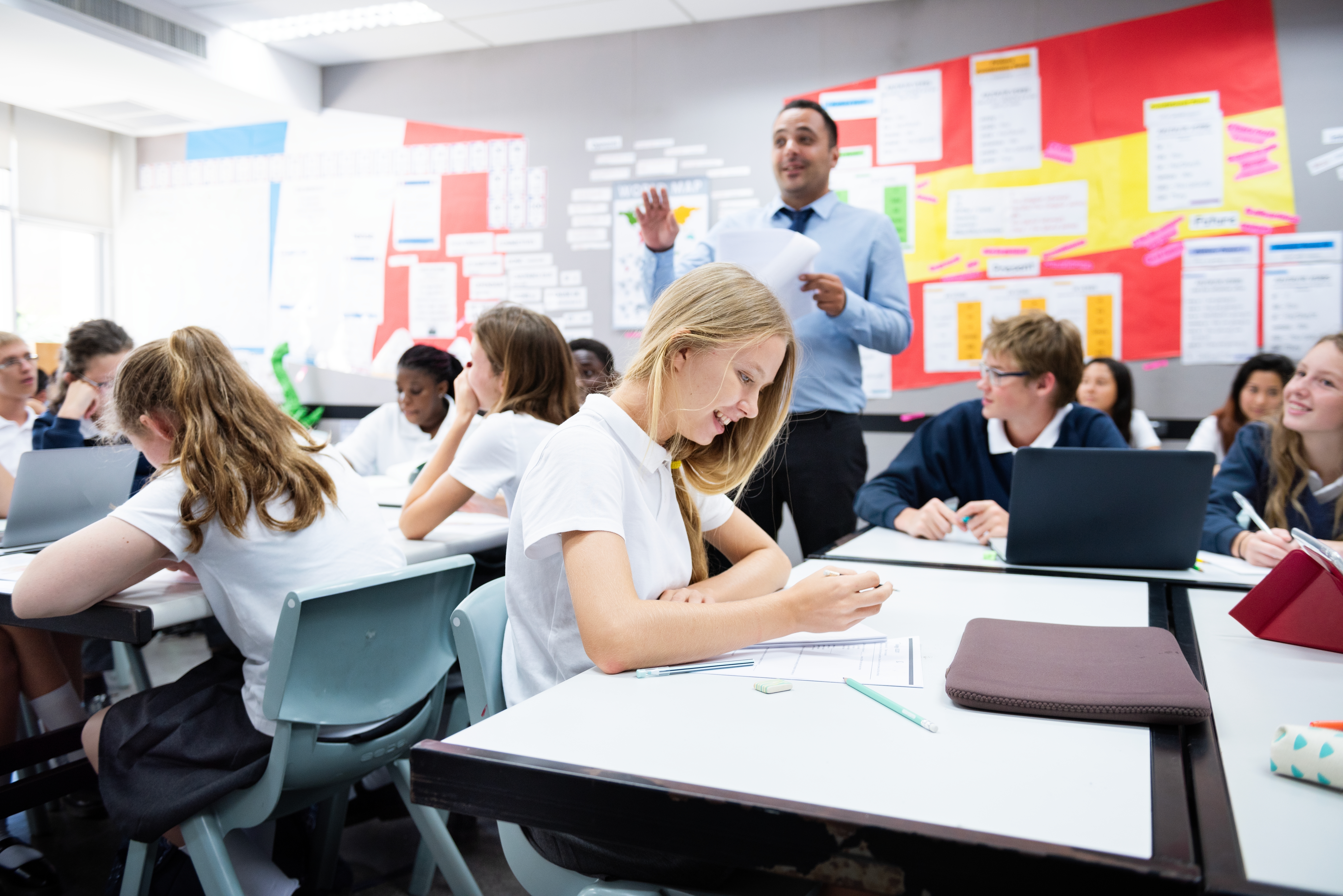 A classroom of secondary school students engaged in a lesson, with a teacher standing at the front speaking while students work at their desks using notebooks and laptops.