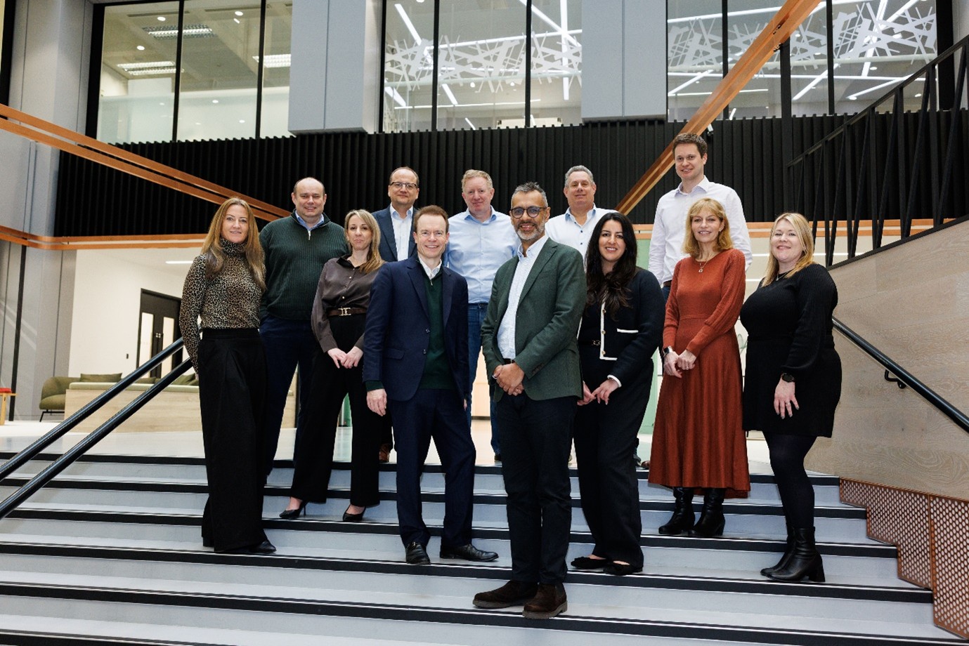 A group of team members from UHY Hacker Young and Affinia's leadership teams, both men and women, stand on indoor stairs in business attire, smiling. The background shows a modern office space with large windows.