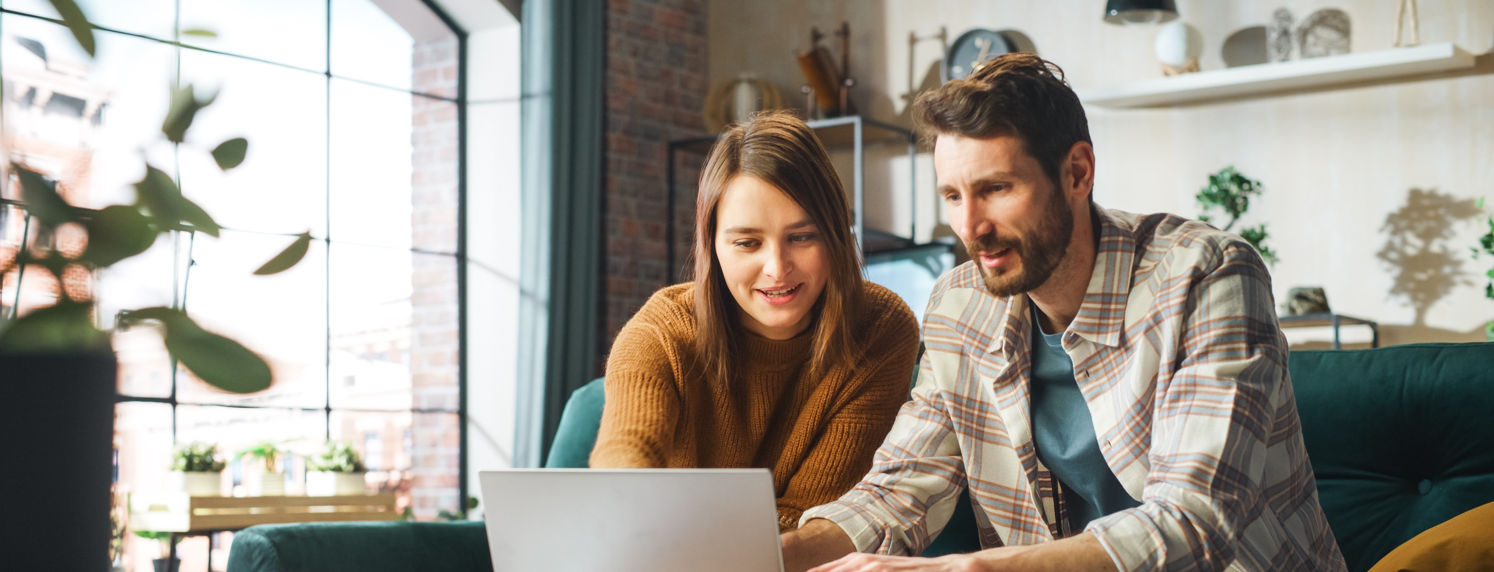 Two people sitting on a sofa in a bright, modern living room, looking at a laptop together and smiling, with papers and shelves in the background.