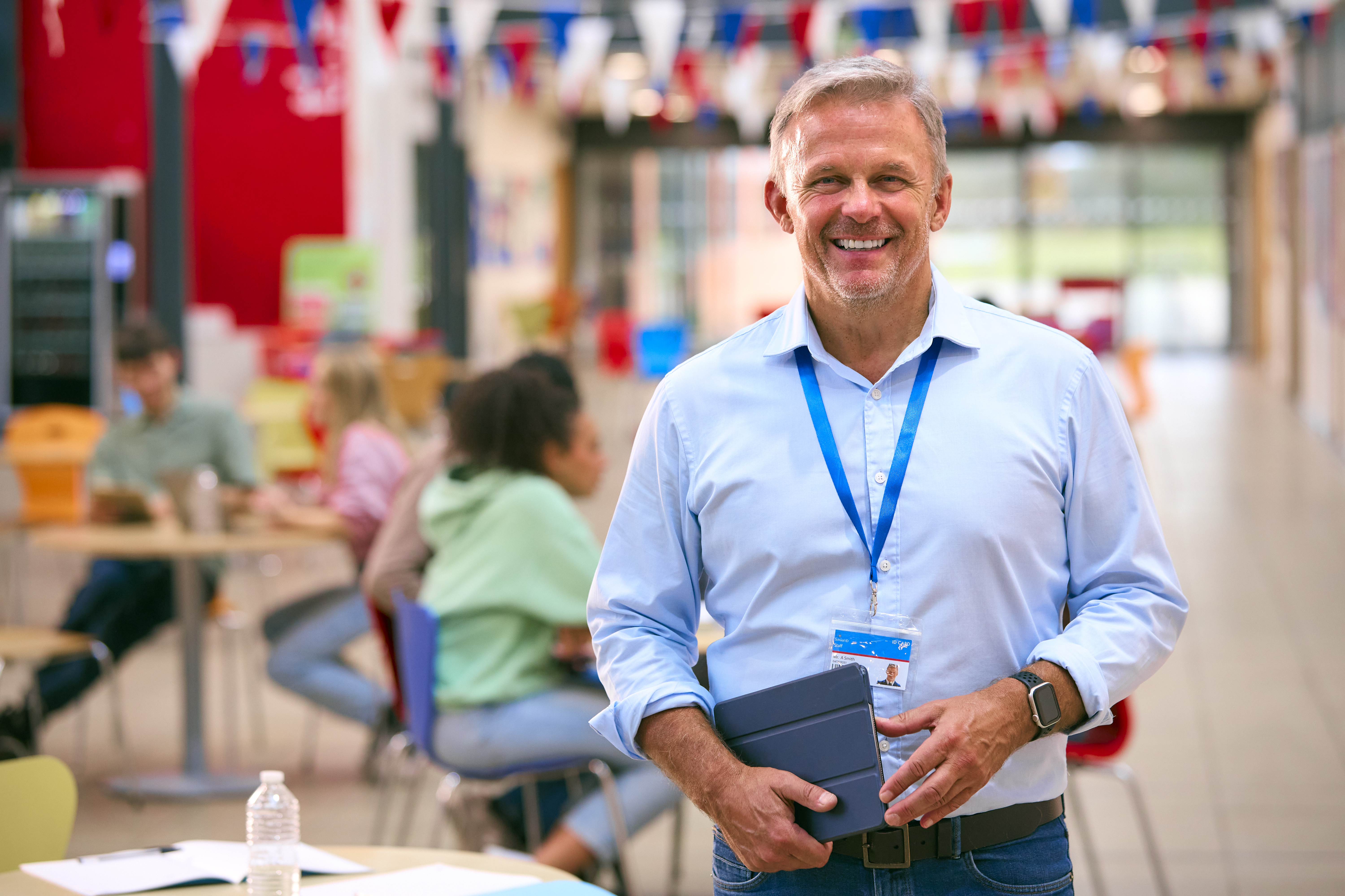 Smiling man with a lanyard holds a tablet in a bright, festive room with blurred people and colorful bunting in the background. Warm and welcoming atmosphere.
