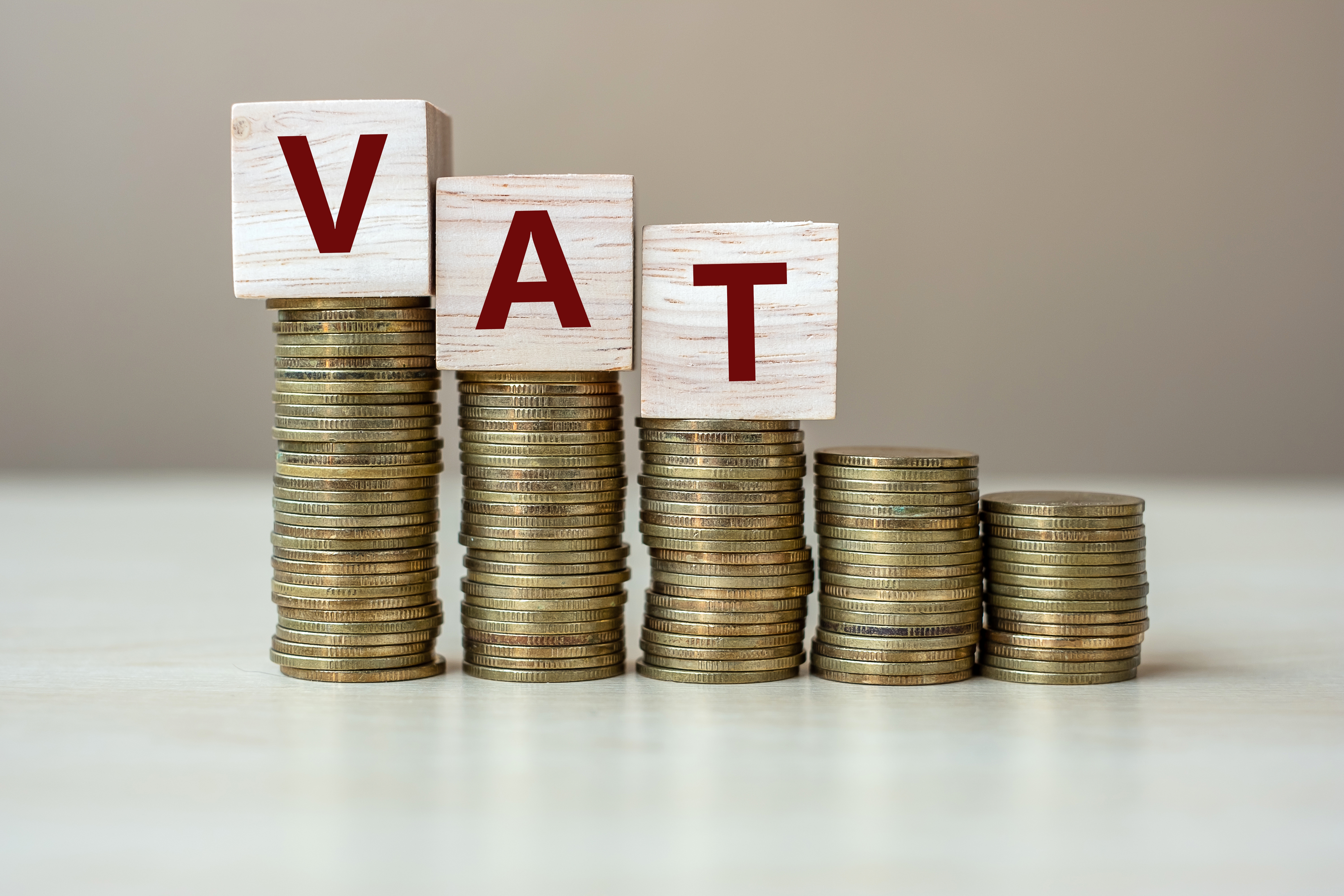 Stacks of coins arranged in varying heights spell out "VAT" using wooden blocks on top. The image symbolizes the financial concept of Value Added Tax.