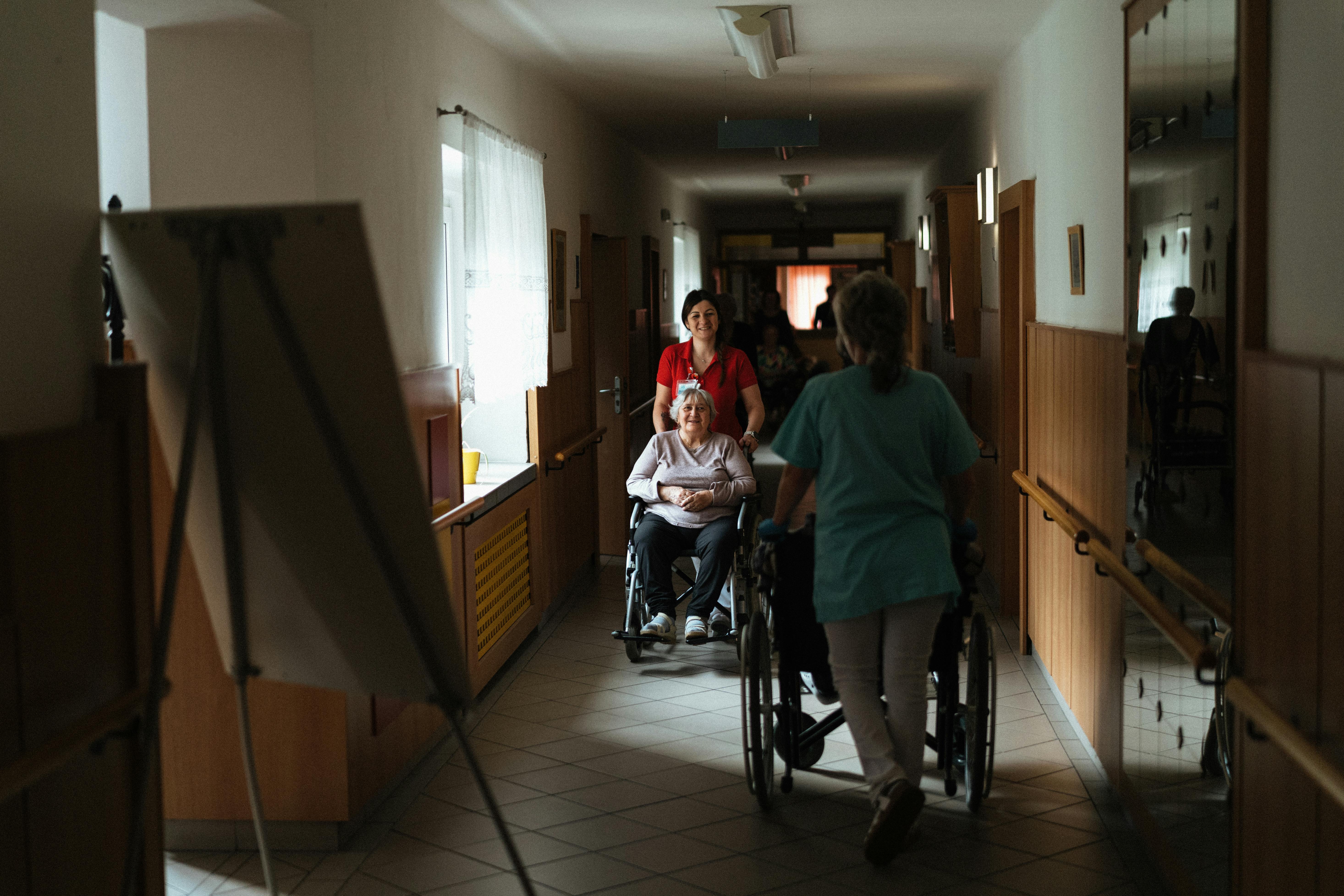 A caregiver in red pushes an elderly woman in a wheelchair down a dimly lit hallway, conveying a calm, caring atmosphere in a nursing home setting.