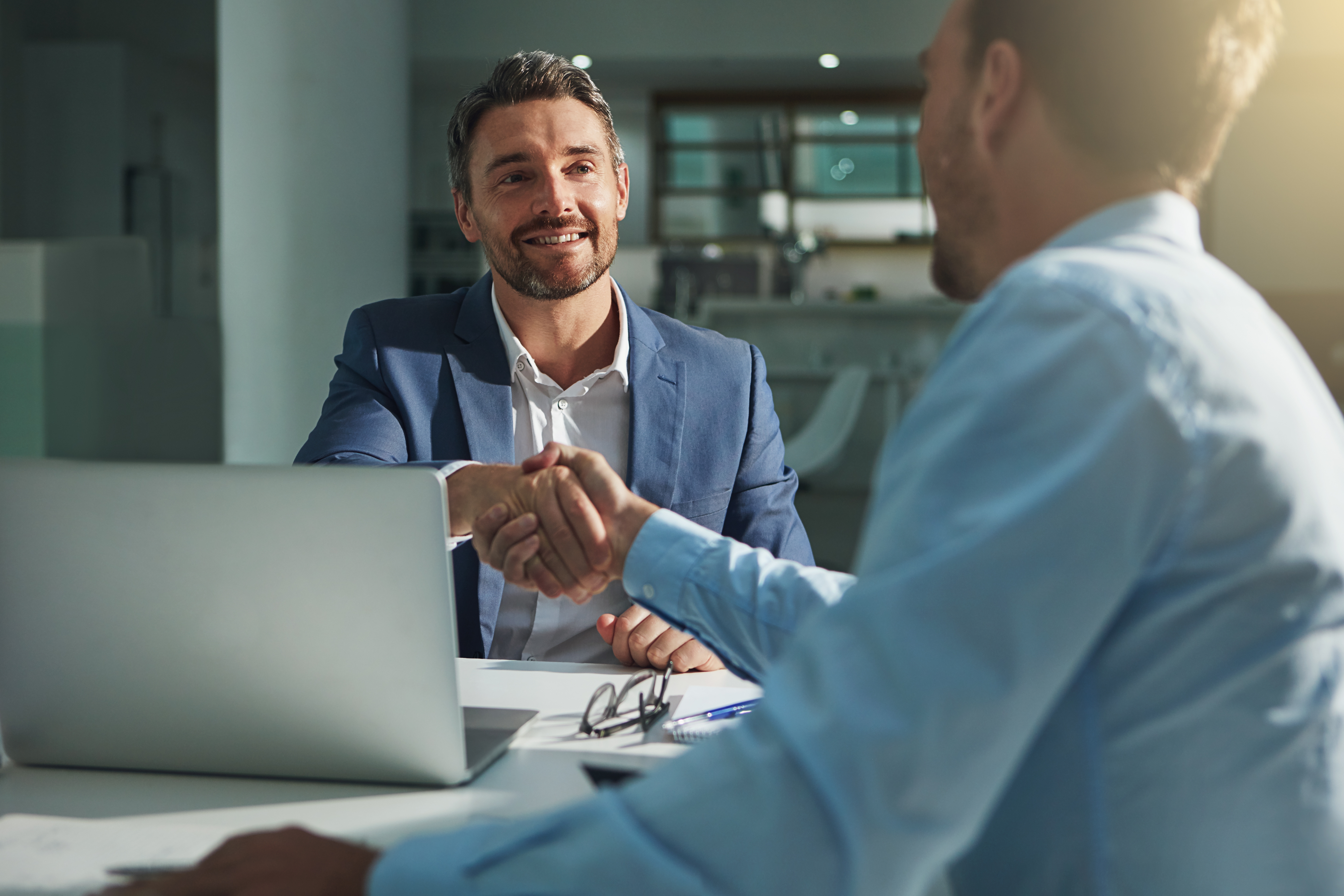 Two men in business attire shake hands across a table in an office setting, with a laptop and glasses beside them, conveying a professional tone.
