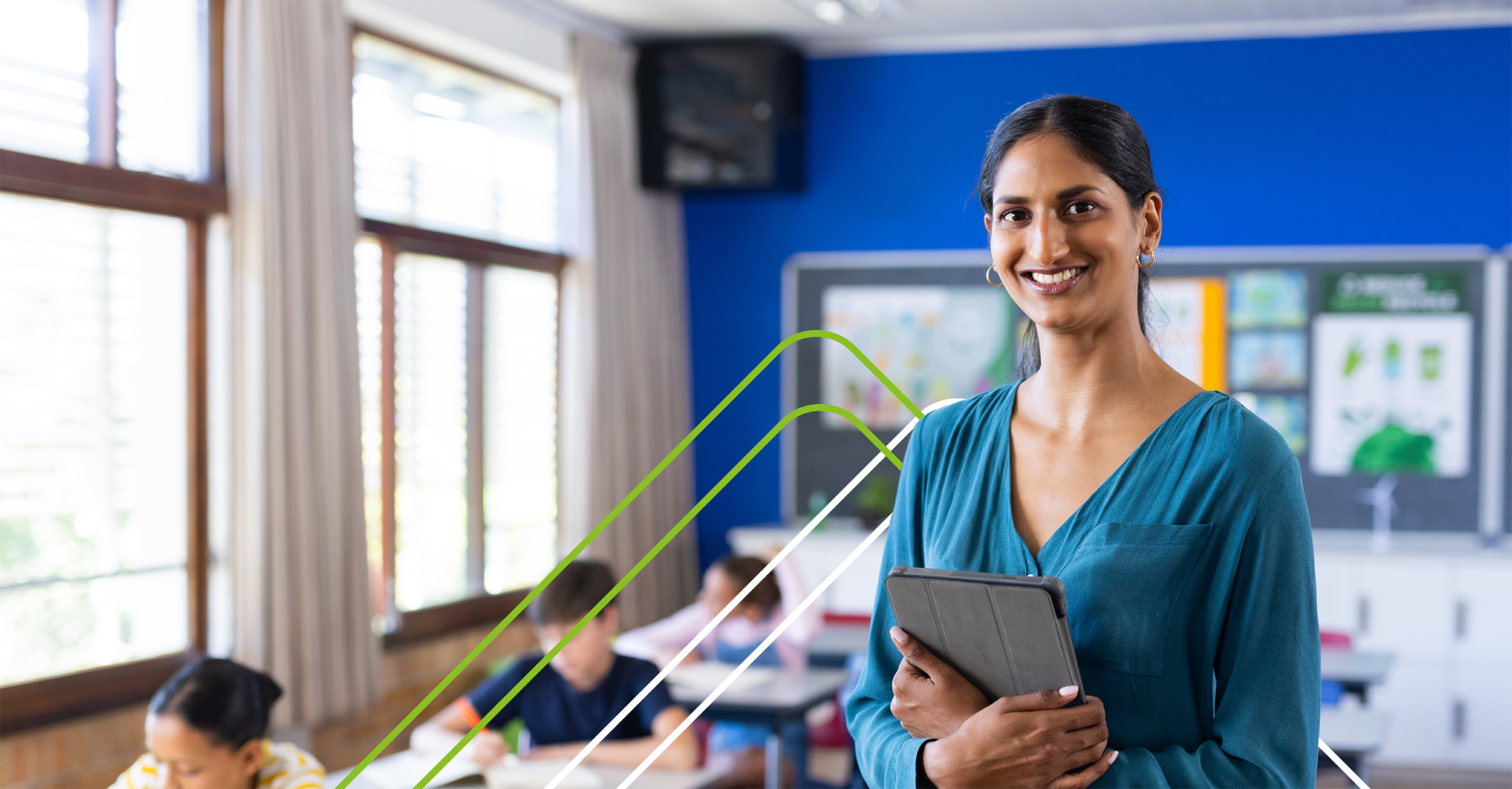 Smiling teacher in a teal blouse holds a tablet in a bright classroom with students working at desks. The atmosphere is positive and engaging.