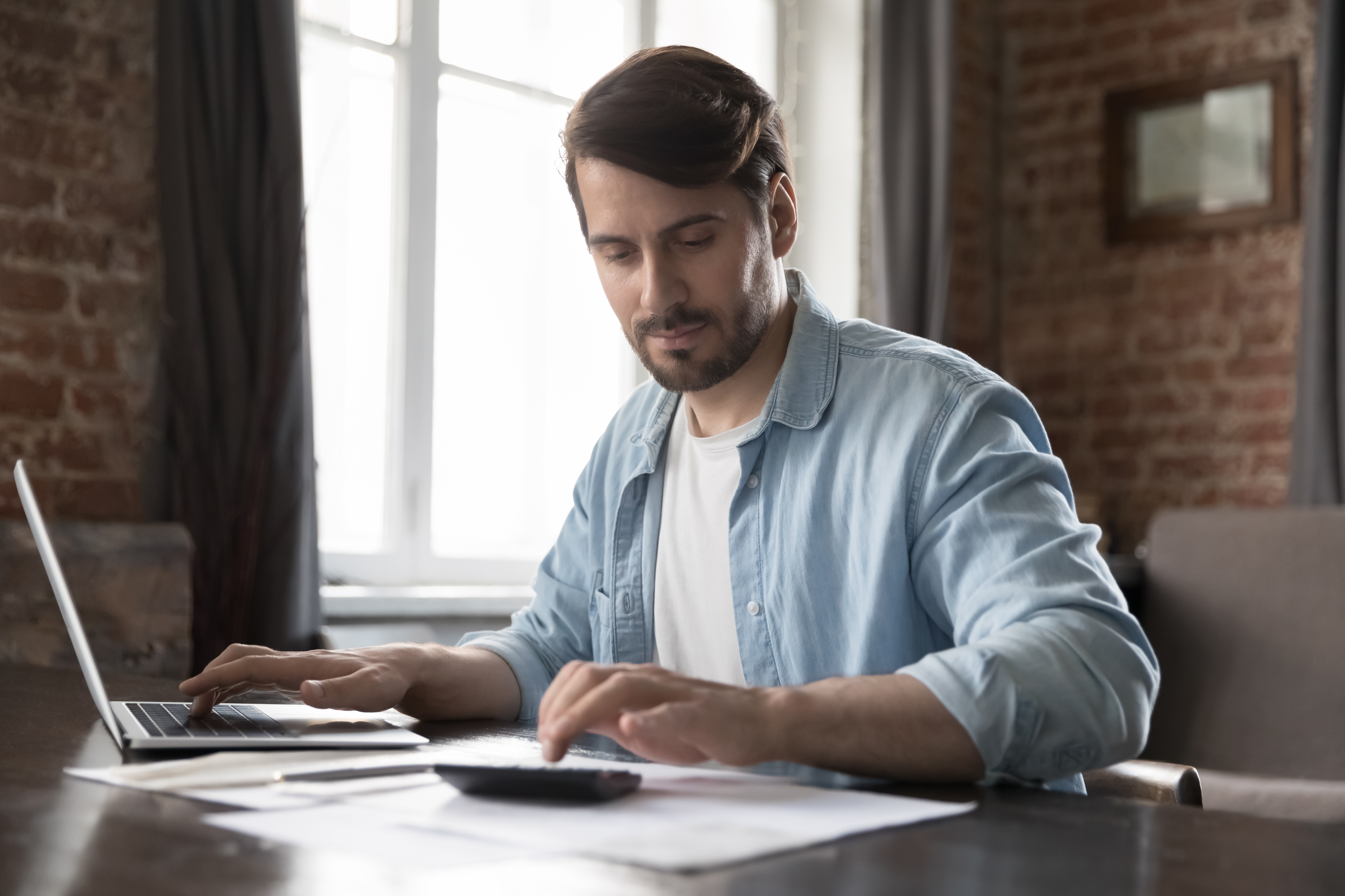 A man in a casual blue shirt works from home, focused on using a calculator and laptop. The scene is well-lit, with a brick wall in the background.