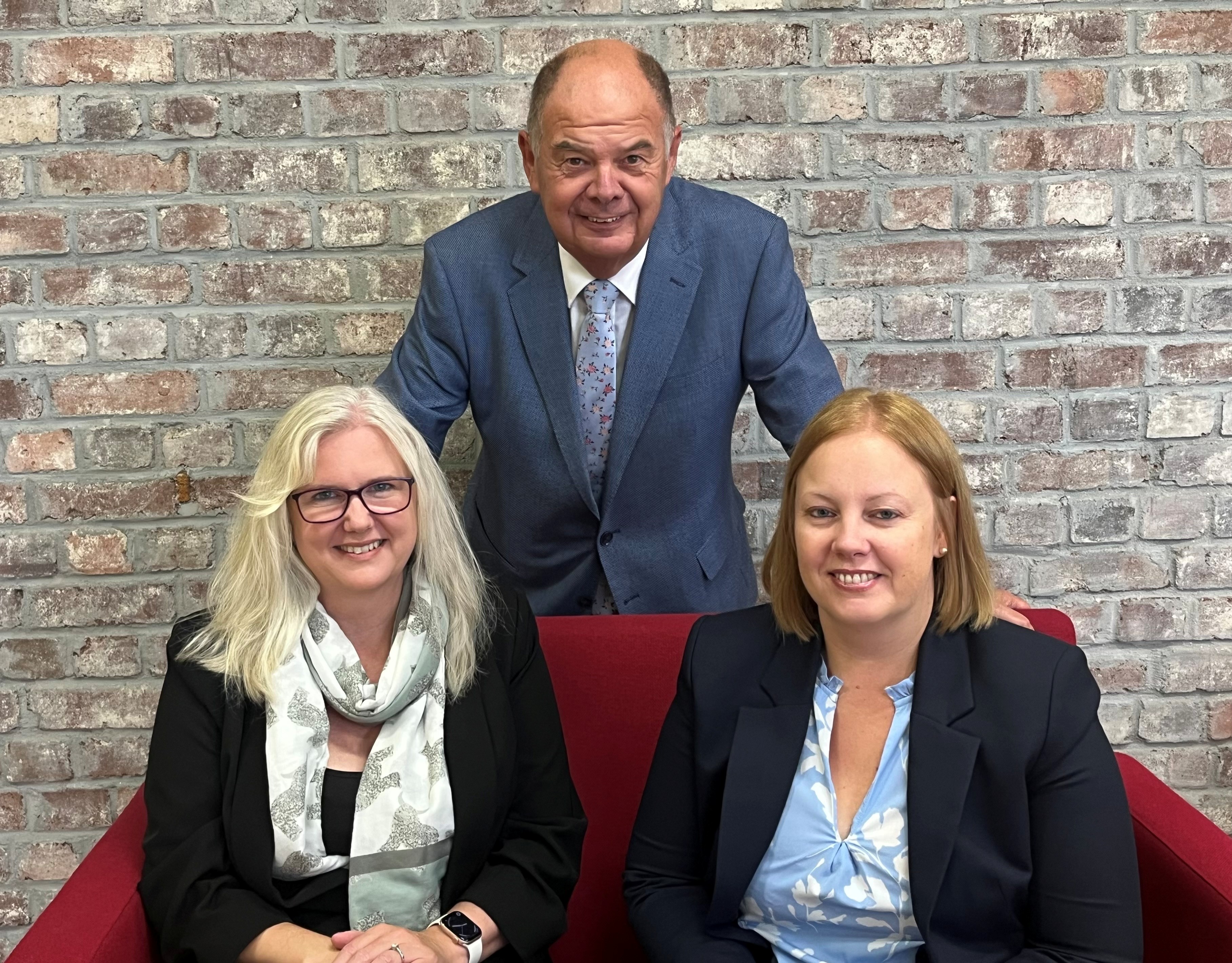 Fiona Davey, Mel Stone and Vince Edwards, partners of UHY Peninsula, smiling indoors against a brick wall. Two women sit on a red couch, while a man stands behind them. All wear business attire.