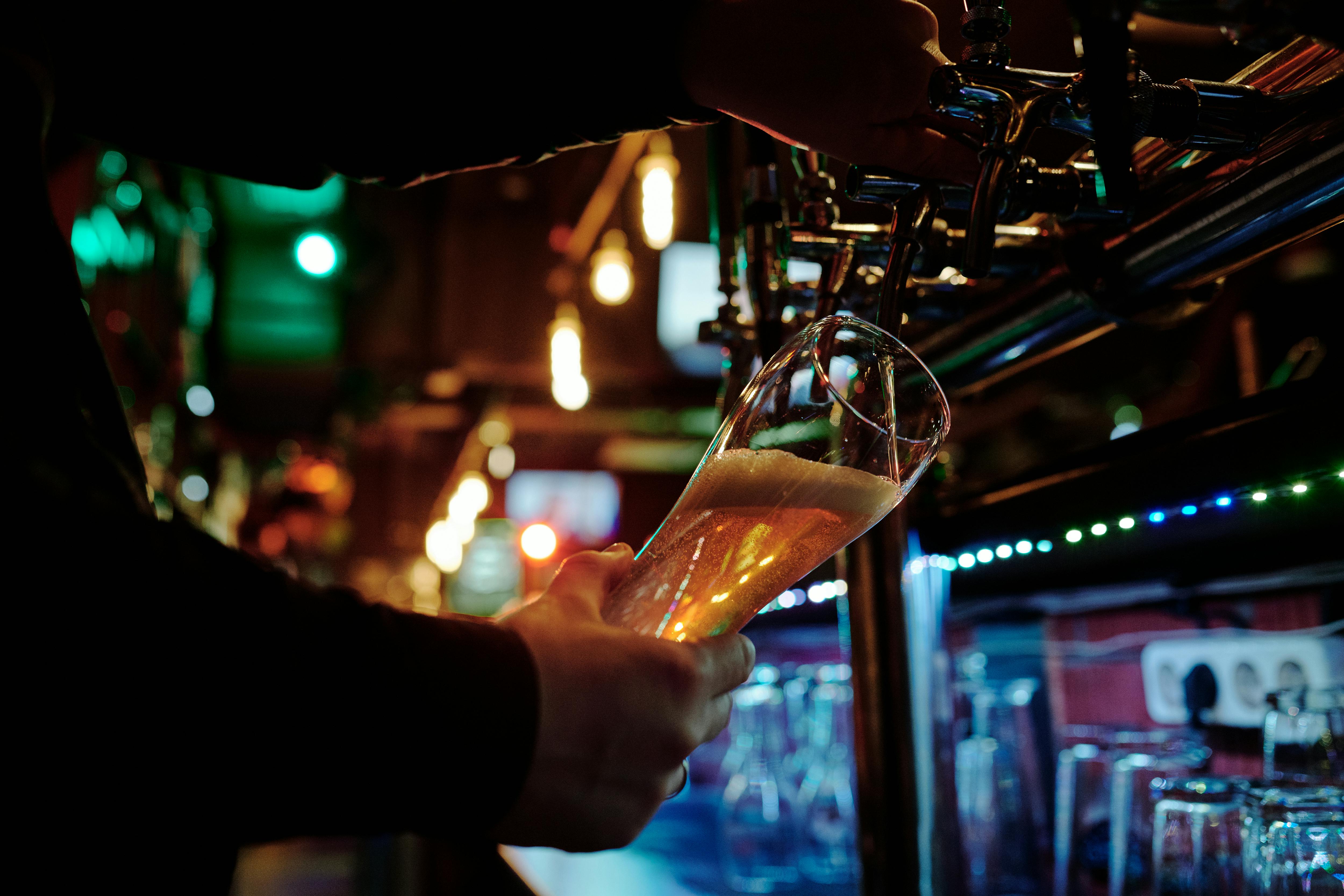 A bartender pours a golden beer into a glass from a tap in a dimly lit bar. Warm lighting and reflections create a cosy, inviting atmosphere.