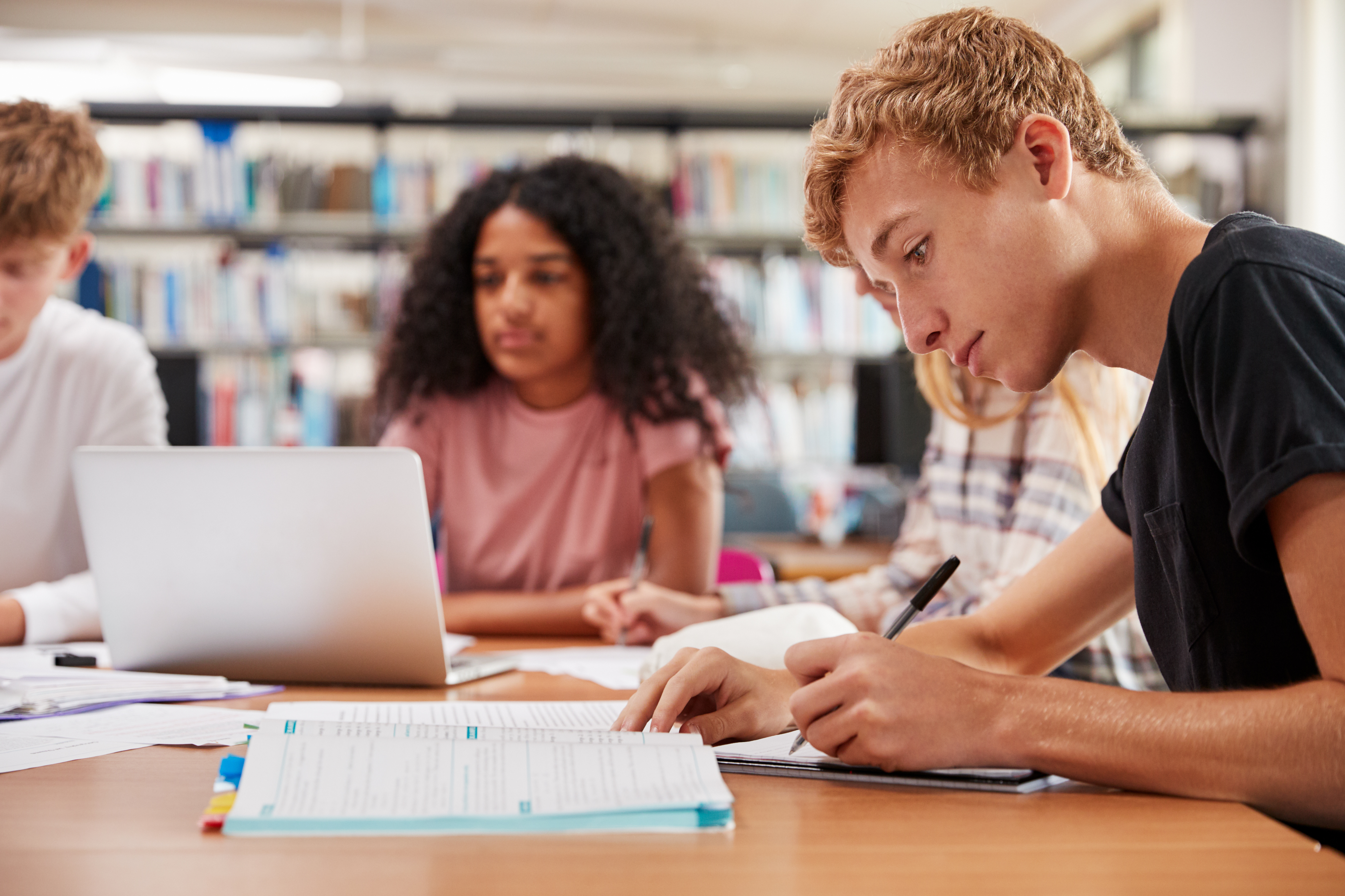 A group of focused students study at a library table. A young man takes notes, while a woman works on a laptop. Shelves of books are in the background.