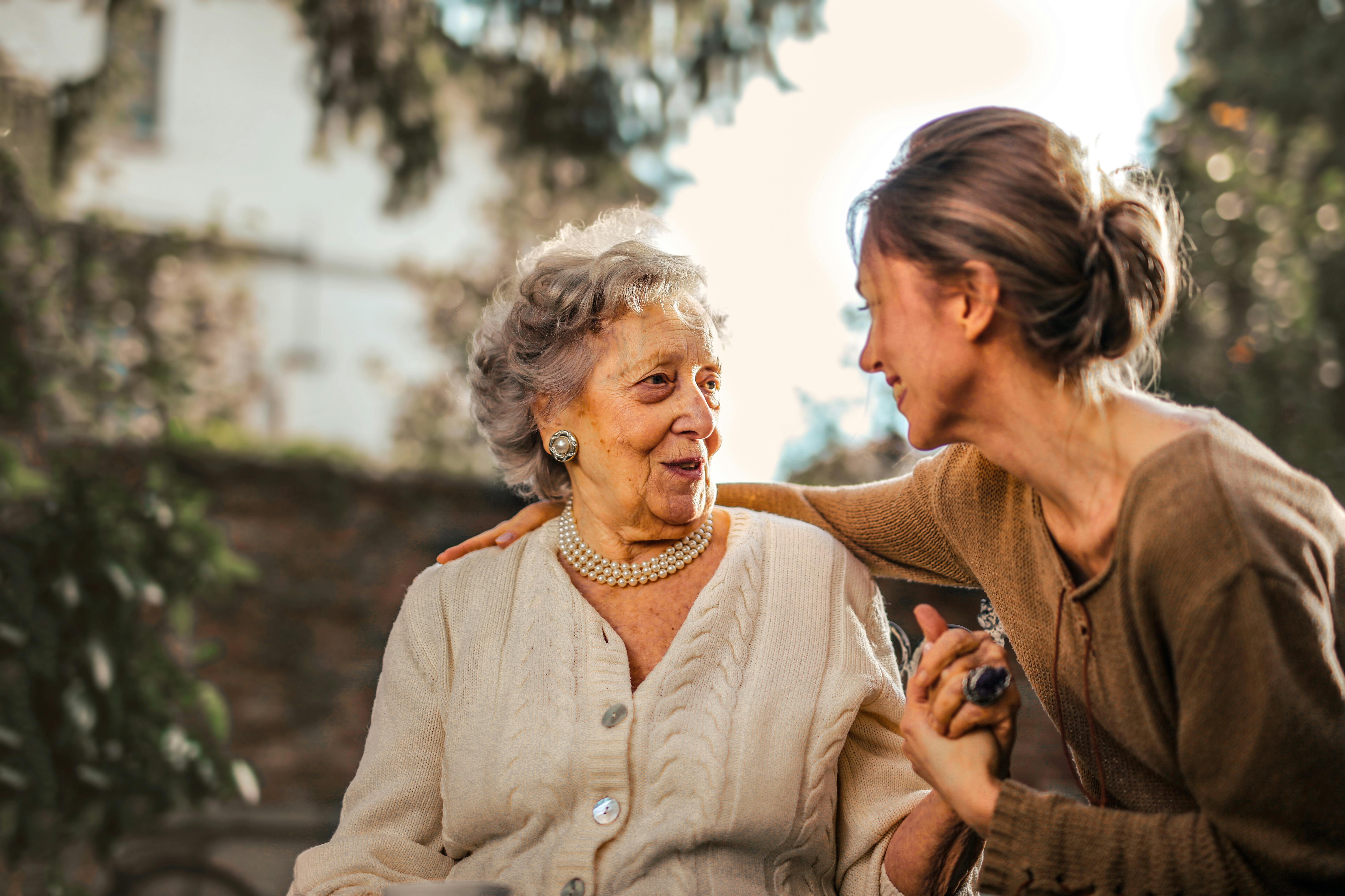 Elderly woman and young woman sitting together outdoors, smiling warmly at each other. The older woman is wearing a pearl necklace and cardigan.