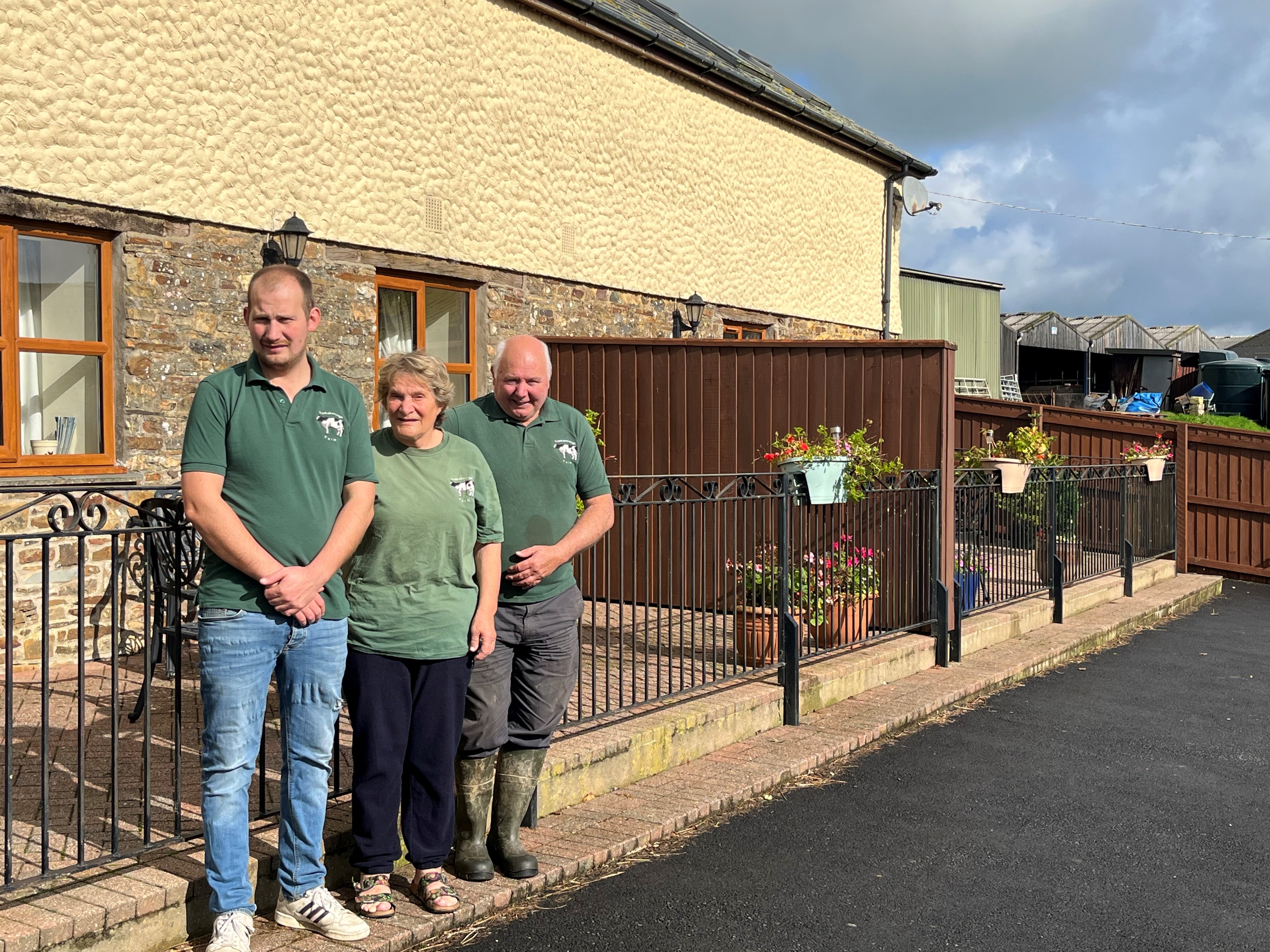 Three people wearing matching green farm-branded polo shirts stand outside a stone farmhouse, next to a railing decorated with hanging flower baskets. Farm buildings and outbuildings are visible in the background under a partly cloudy sky.