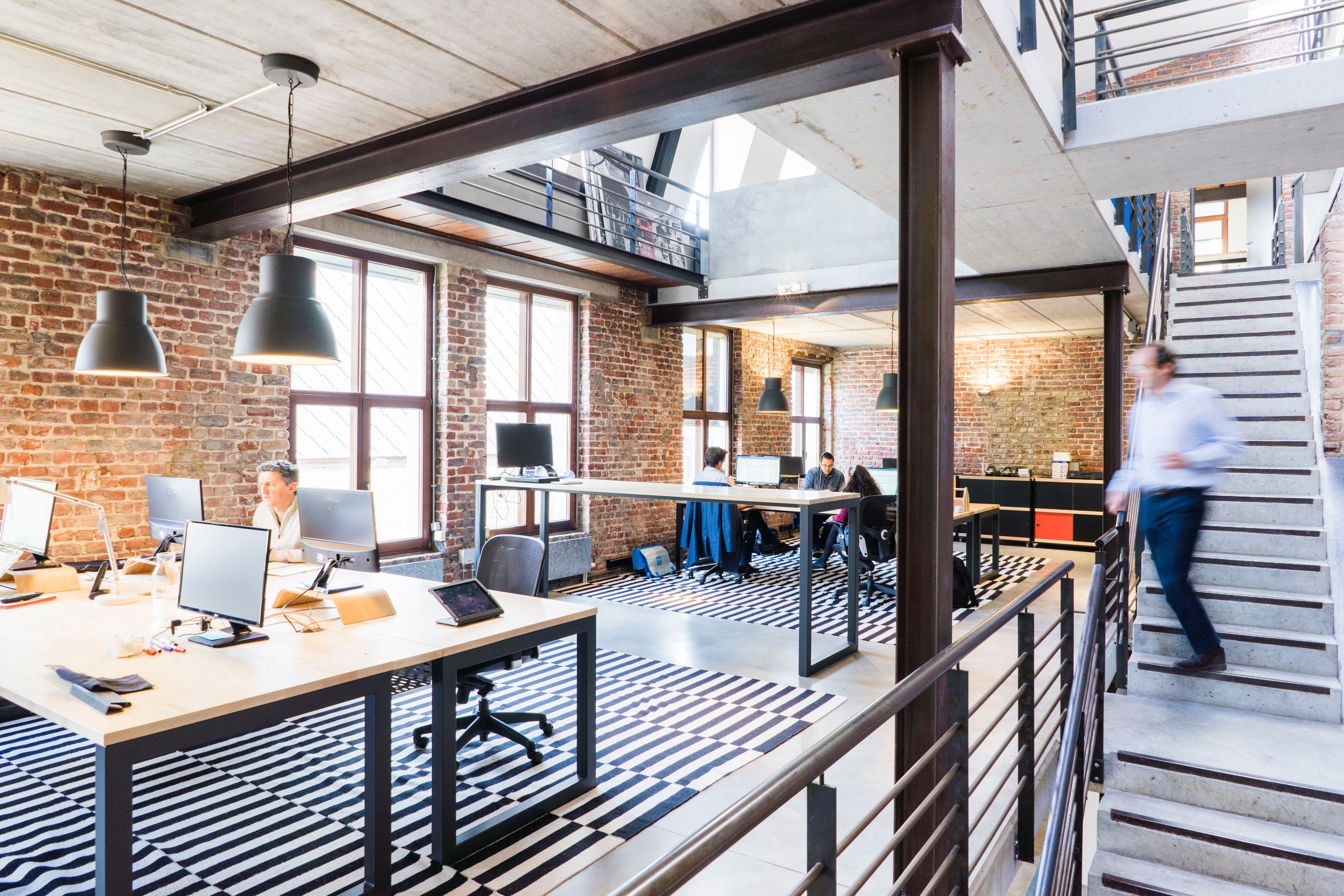 Modern open-plan office with exposed brick walls, steel beams and a mezzanine level. Several people work at desks with computer monitors, while a blurred figure walks up a concrete staircase on the right. Large windows provide natural light and black pendant lamps hang above the workspace. Black-and-white striped rugs define the seating areas.