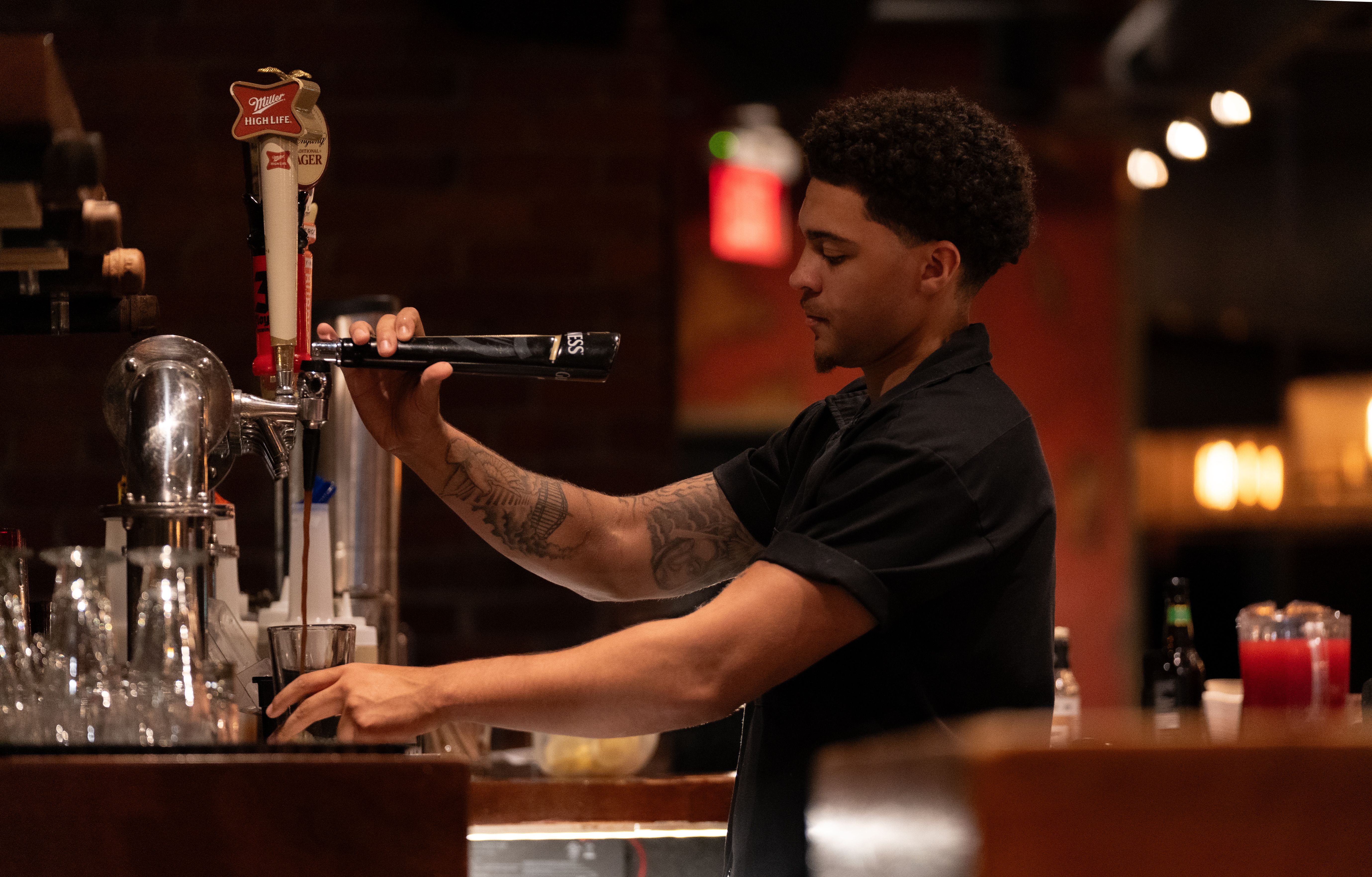 A person behind a bar pours a drink from a beer tap into a glass. Rows of clean glasses sit on the counter in the foreground, while warm ambient lighting and a softly blurred restaurant or bar interior create a cosy atmosphere in the background. Visible branding on the tap includes Miller High Life and Guinness.