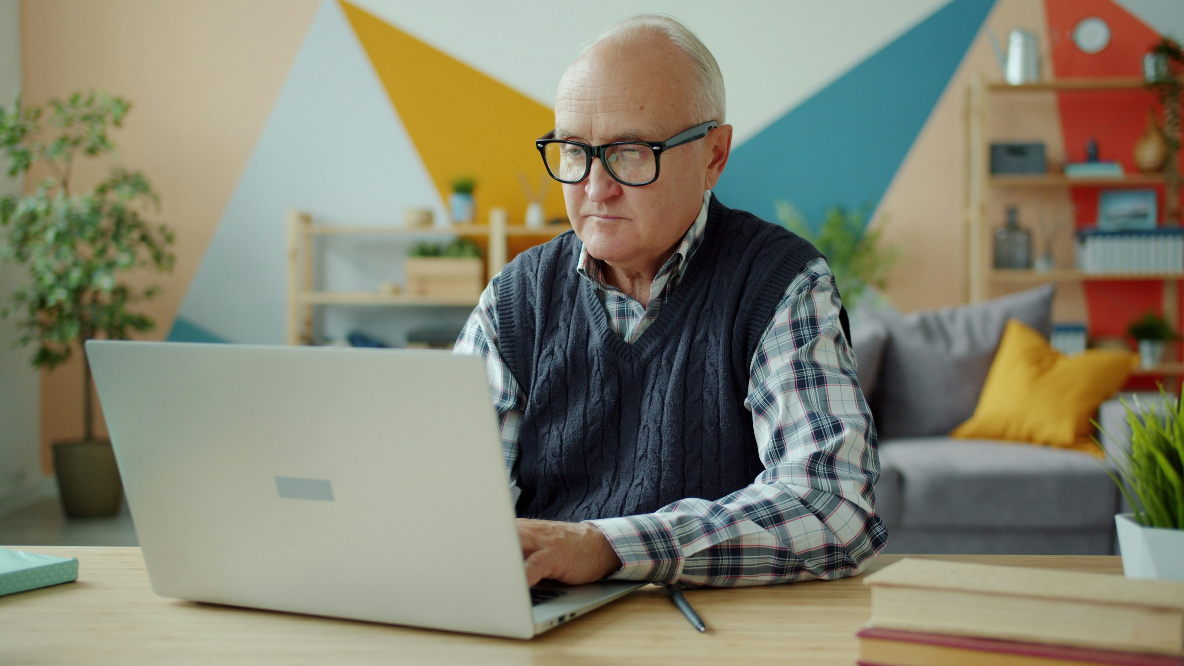 Elderly man with glasses focused on a laptop at a wooden desk. The bright, geometric wall and cozy decor create a modern, comfortable atmosphere.