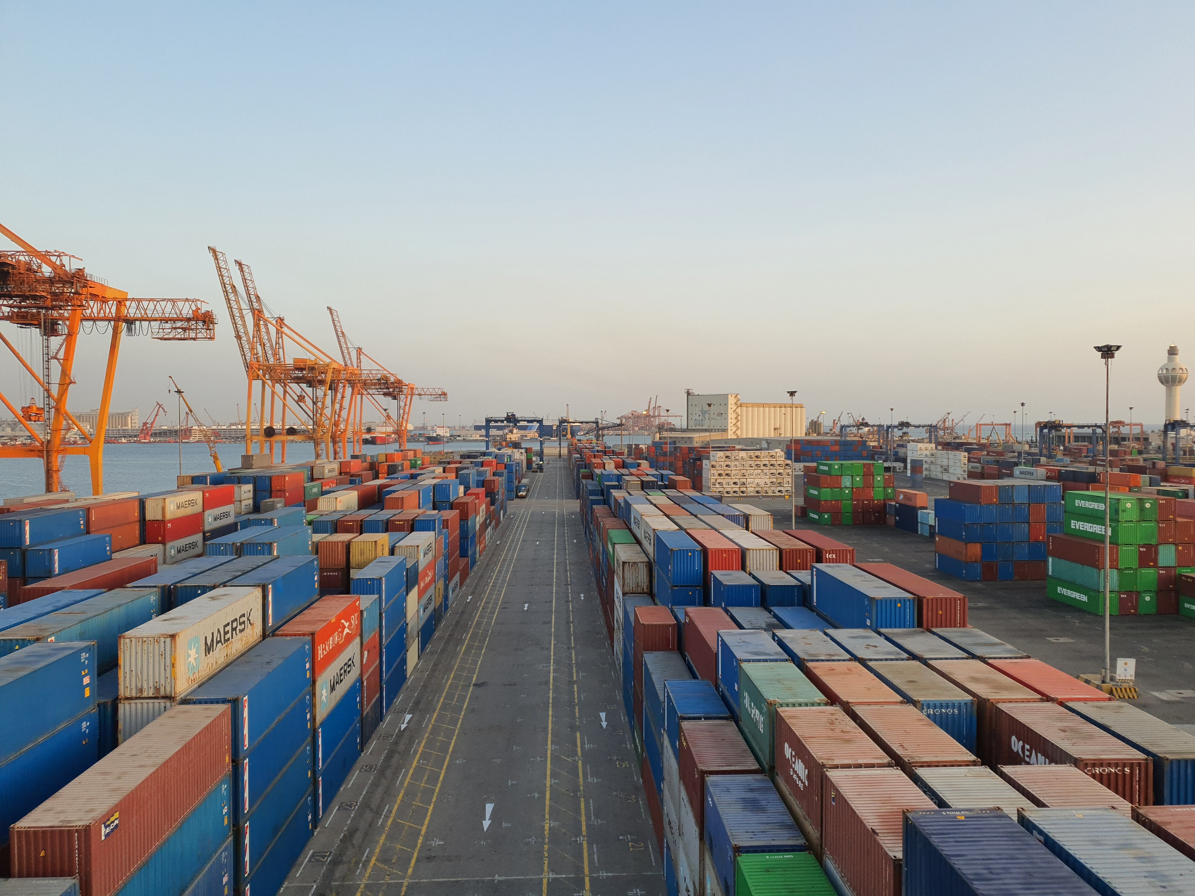 Elevated view of a busy container port at dusk, with long rows of stacked shipping containers from lines and tall orange gantry cranes on the waterfront to the left.