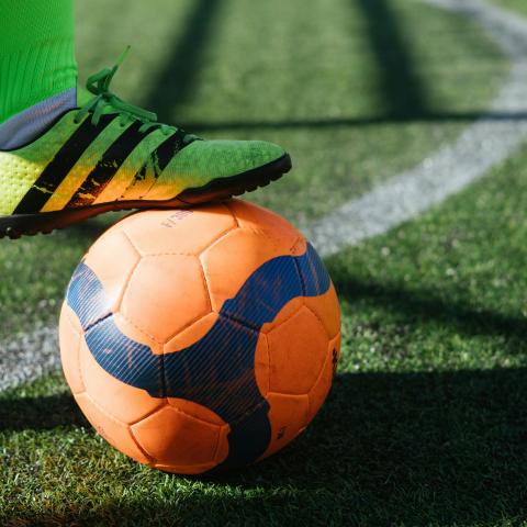 footballer with neon socks and football trainers, standing on a football pitch, with a football held under their foot.