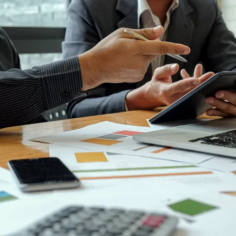 hands of two accountants pointing at calculator with a laptop in front of them and lots of paperwork around them on the table