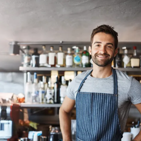 happy business owner in a bar smiling at camera, with drinks behind him on a shelf
