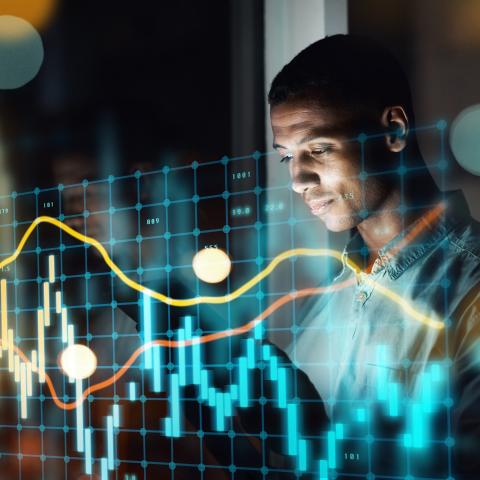 A man standing in a dark room, looking at a digital screen displaying complex financial data and graphs. The screen overlays various line charts, bar graphs, and numerical data, illuminated with bright colors like blue, yellow, and red, suggesting data analysis or financial market monitoring.
