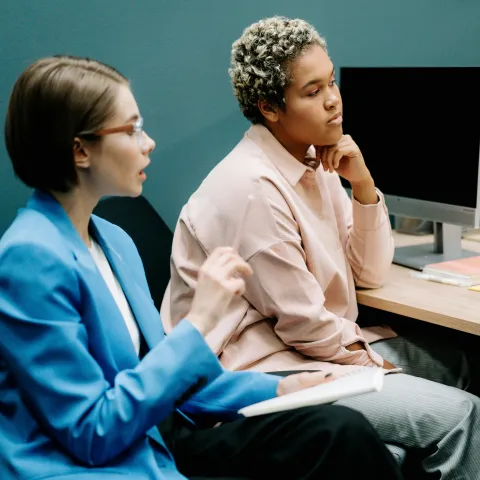 Two individuals are seated in an office setting. The person on the left, wearing glasses and a bright blue blazer, is holding a pen and appears to be speaking or explaining something. The person on the right, dressed in a light pink shirt, is listening attentively with their hand resting on their chin. Both are focused, with a computer monitor and notebooks on the desk in front of them.