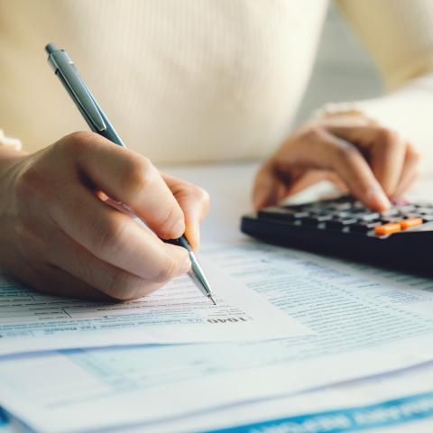 A person in a light-colored sweater using a calculator while writing on financial documents, indicating tax or financial calculations.