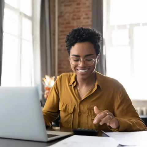 Happy young Afro American entrepreneur woman in glasses counting profit, on calculator at laptop computer, analyzing benefits, enjoying financial success, job high result, smiling