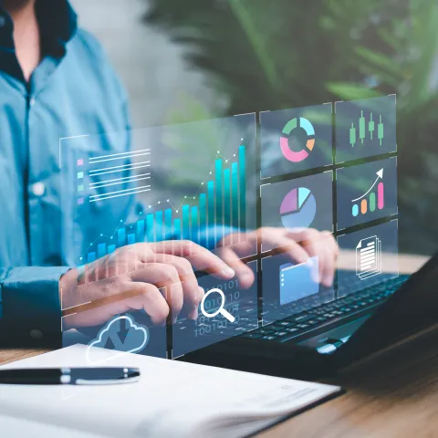 A person in a blue shirt types on a laptop at a desk, with a notebook and pen nearby. Overlaid digital graphics display data visualizations, including bar charts, pie charts, and cloud storage icons, representing financial or business analytics. The background features greenery and natural light, creating a modern, tech-focused workspace.