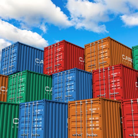 Stacked cargo shipping containers in various colours (blue, red, green, and orange) under a bright blue sky with scattered clouds, symbolising global trade and logistics.