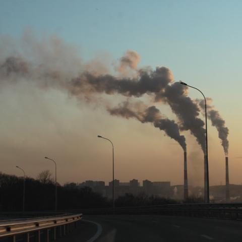 Industrial smokestacks emitting thick plumes of smoke into the sky at sunset, with a city skyline and an empty curved highway in the foreground, highlighting air pollution and environmental impact.