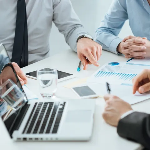 Three business professionals in formal attire are seated around a table discussing financial documents with graphs and charts. A laptop, tablet, smartphone, and glasses of water are also on the table, indicating a collaborative office meeting.