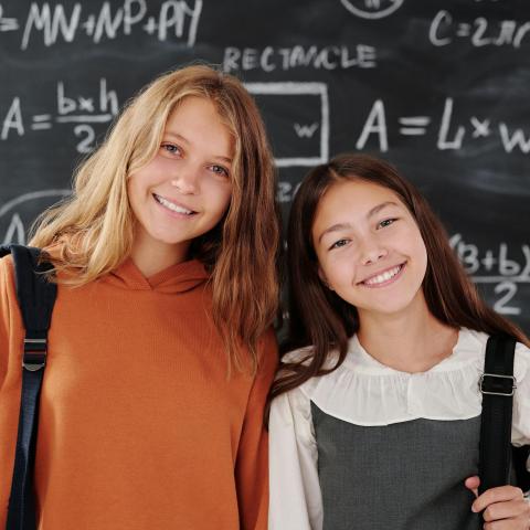 Two smiling schoolgirls stand in front of a blackboard covered with math formulas and diagrams. One wears an orange hoodie, the other a white blouse with a grey pinafore. Both have backpacks over their shoulders.