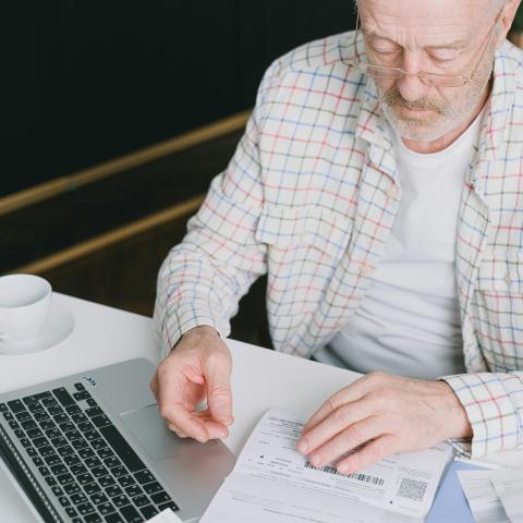 Older man wearing glasses and a plaid shirt sitting at a white table, reviewing documents next to an open laptop. A cup and saucer are placed nearby.