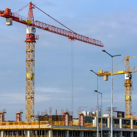 Construction site with multiple yellow and red tower cranes operating over a multi-storey building framework under development, against a blue sky.