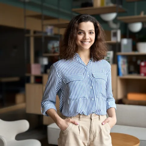 Young woman standing confidently in a modern office lounge area, smiling at the camera. She is wearing a blue and white striped blouse with beige trousers, with her hands in her pockets. The background features shelves, books, and modern furniture in a relaxed, contemporary setting.