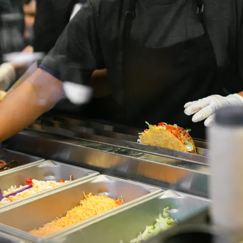 A food service worker wearing gloves and a black apron prepares a taco behind a counter with various fresh ingredients including shredded cheese, lettuce, and salsa.