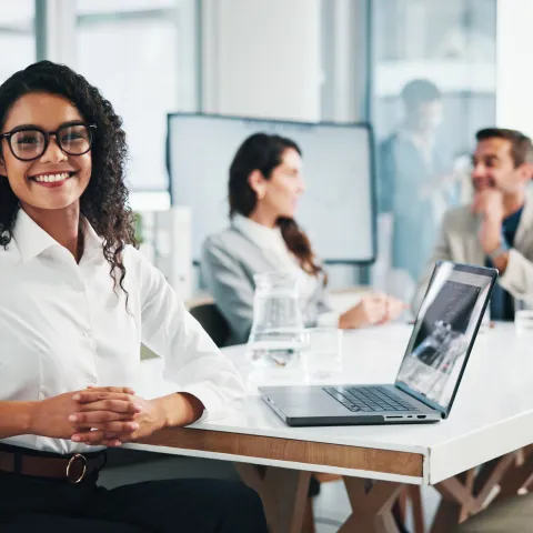 A woman sits at a desk with a laptop, smiling at the camera, while two colleagues have a discussion in the background in a modern office setting.