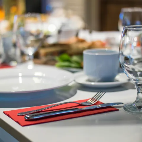 Close-up of a dining table set with white plates, glassware, a coffee cup, and silver cutlery placed on a red napkin.