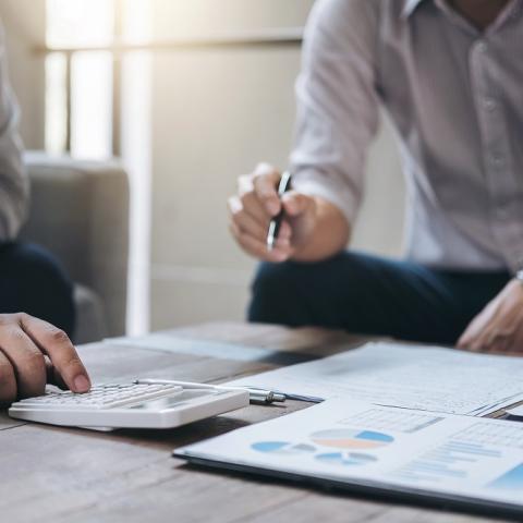 Two professionals reviewing financial documents and charts on a table, with one person using a calculator and the other holding a pen during a business discussion.