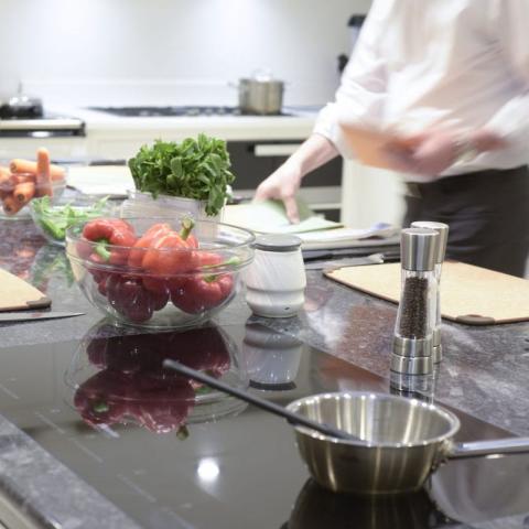 A modern kitchen workspace with fresh vegetables, herbs, and cooking utensils on a countertop, while a chef prepares food in the background.